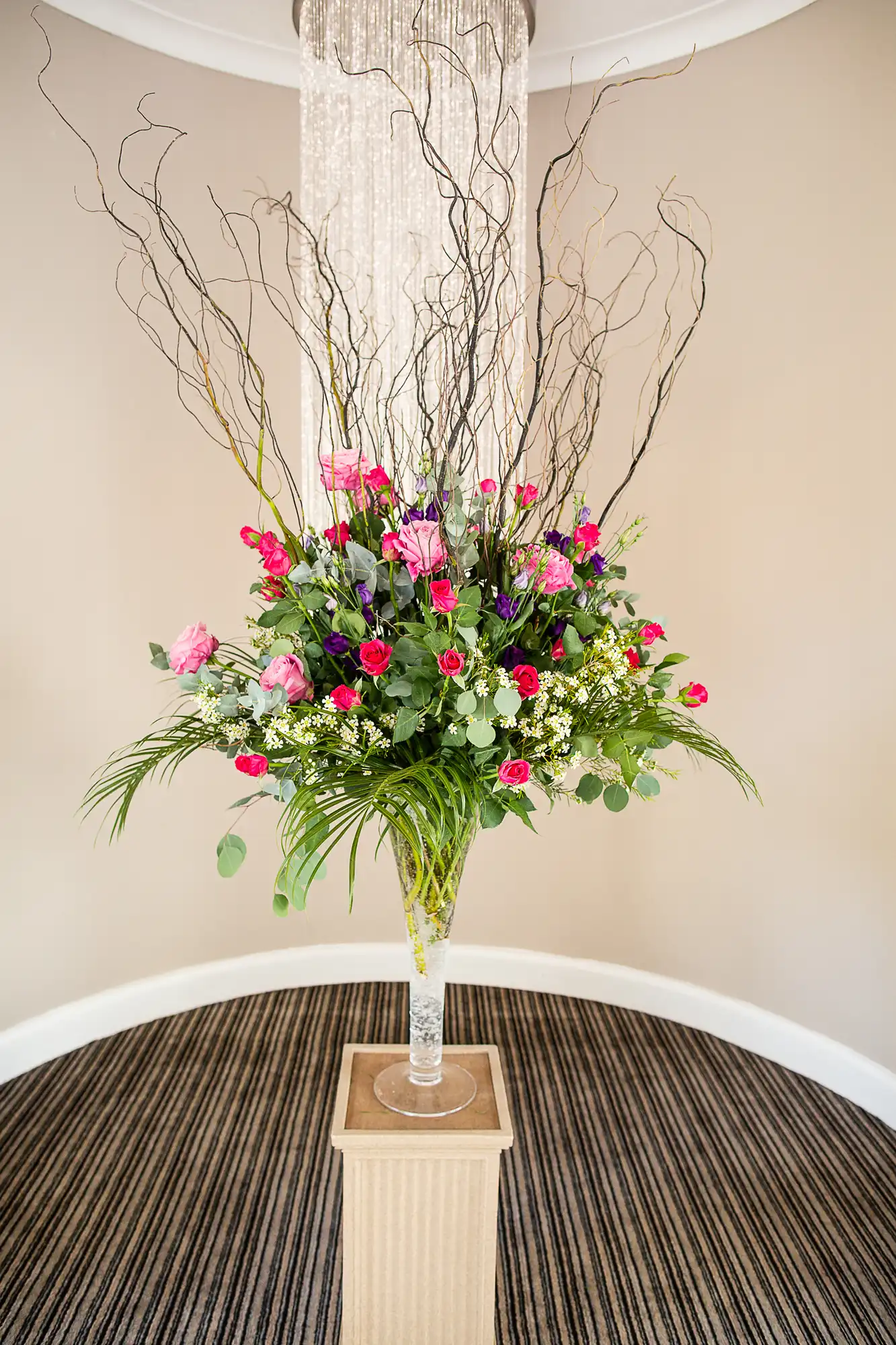 Elegant floral arrangement with pink roses and green foliage in a tall glass vase on a pedestal, under a crystal chandelier.