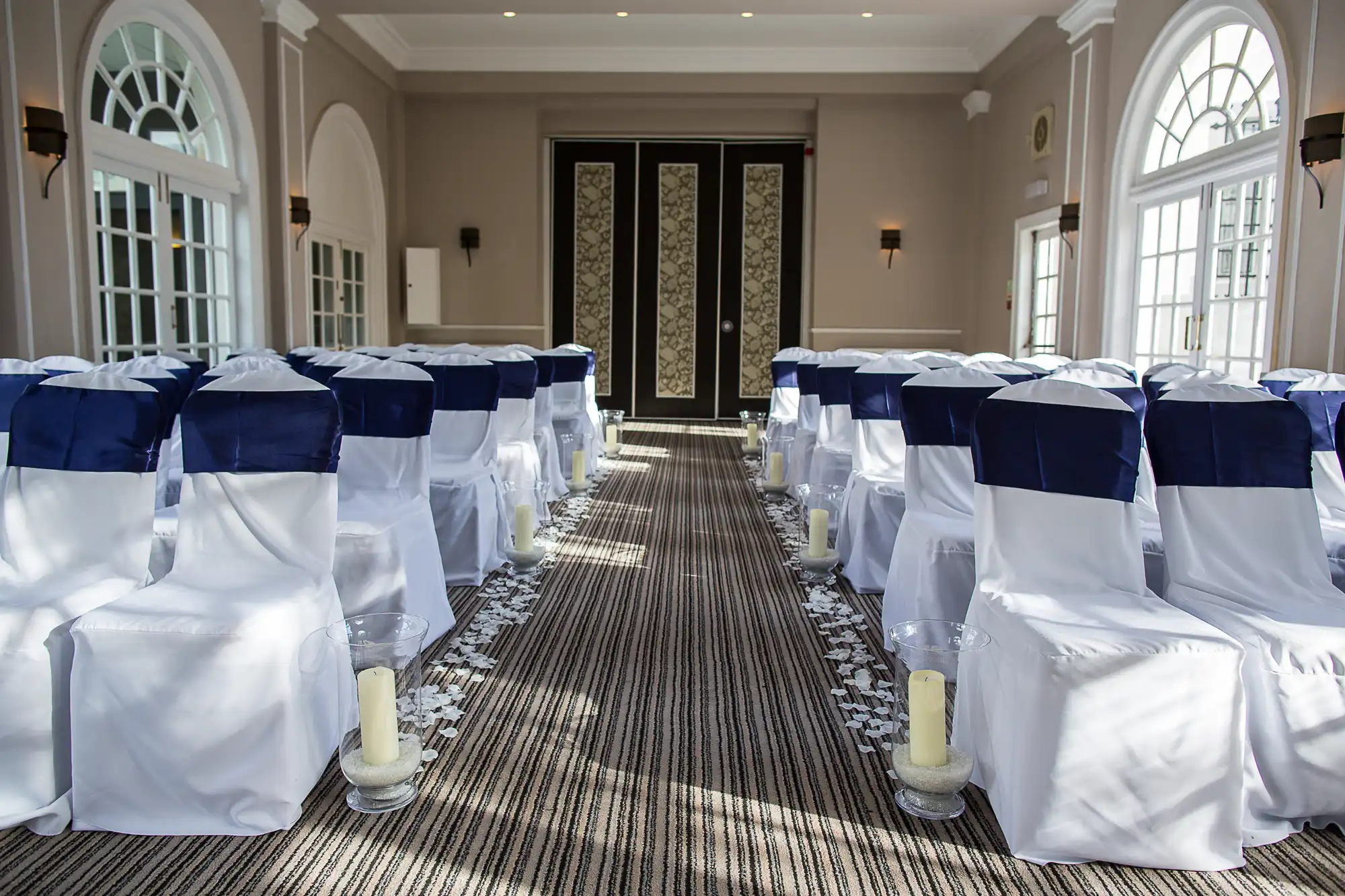 Elegant wedding venue interior with rows of white chairs adorned with dark blue sashes, a patterned aisle runner, and floor candles, illuminated by natural light from large windows.