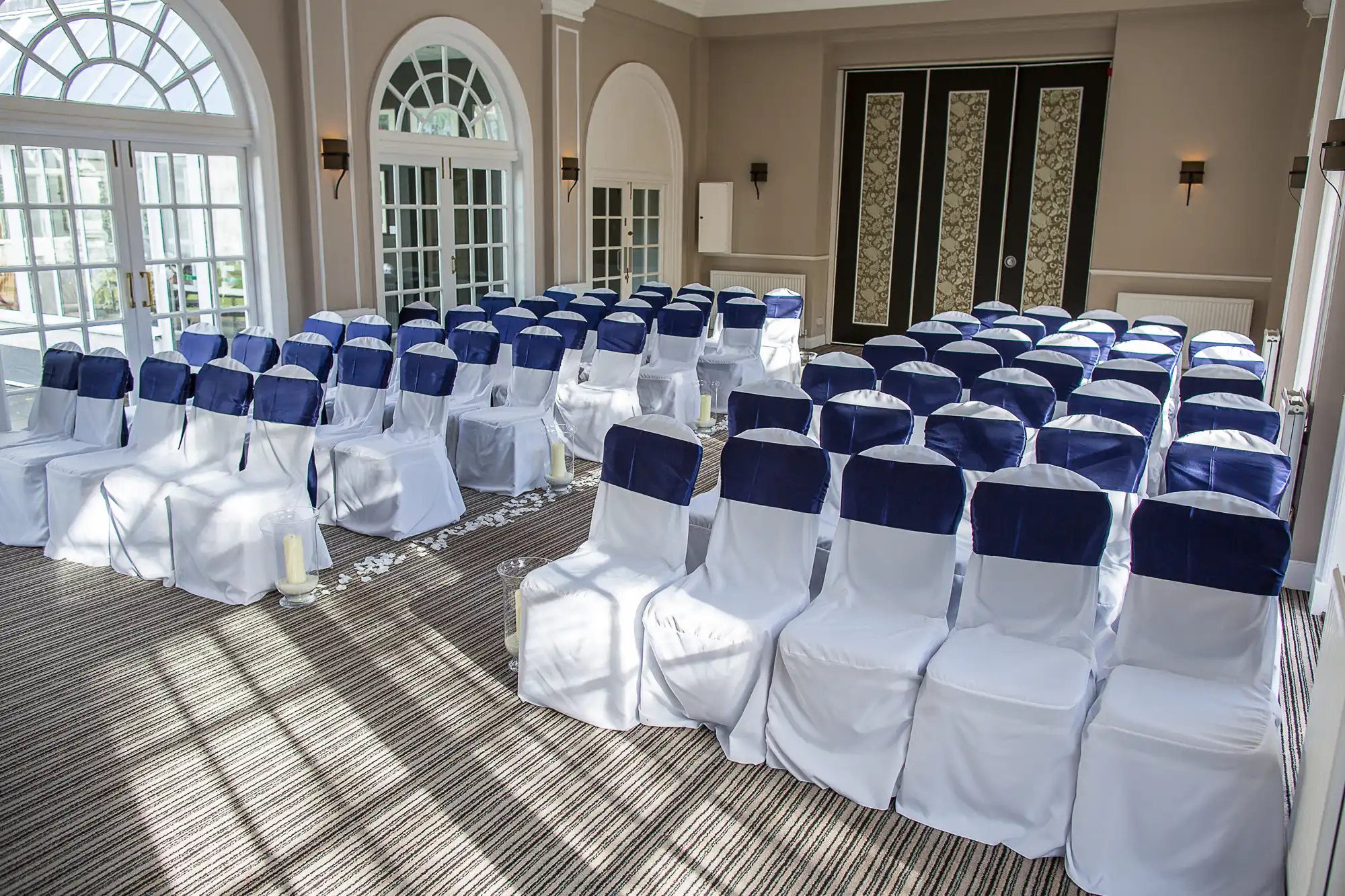 Elegant event venue with rows of white chairs covered in blue sashes, arranged on a striped carpet, facing large arched windows.