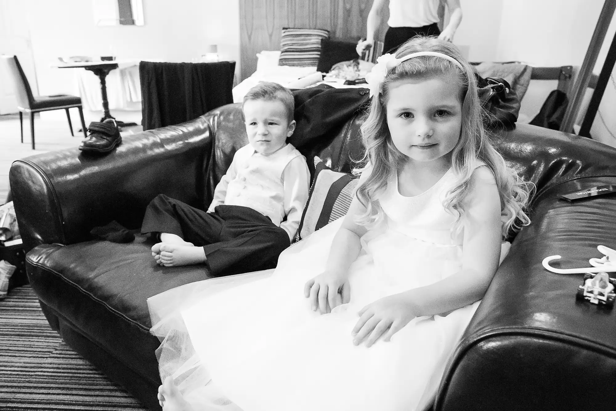 A young girl in a white dress and a young boy in a white shirt and black pants, sitting on a black leather couch, looking at the camera.
