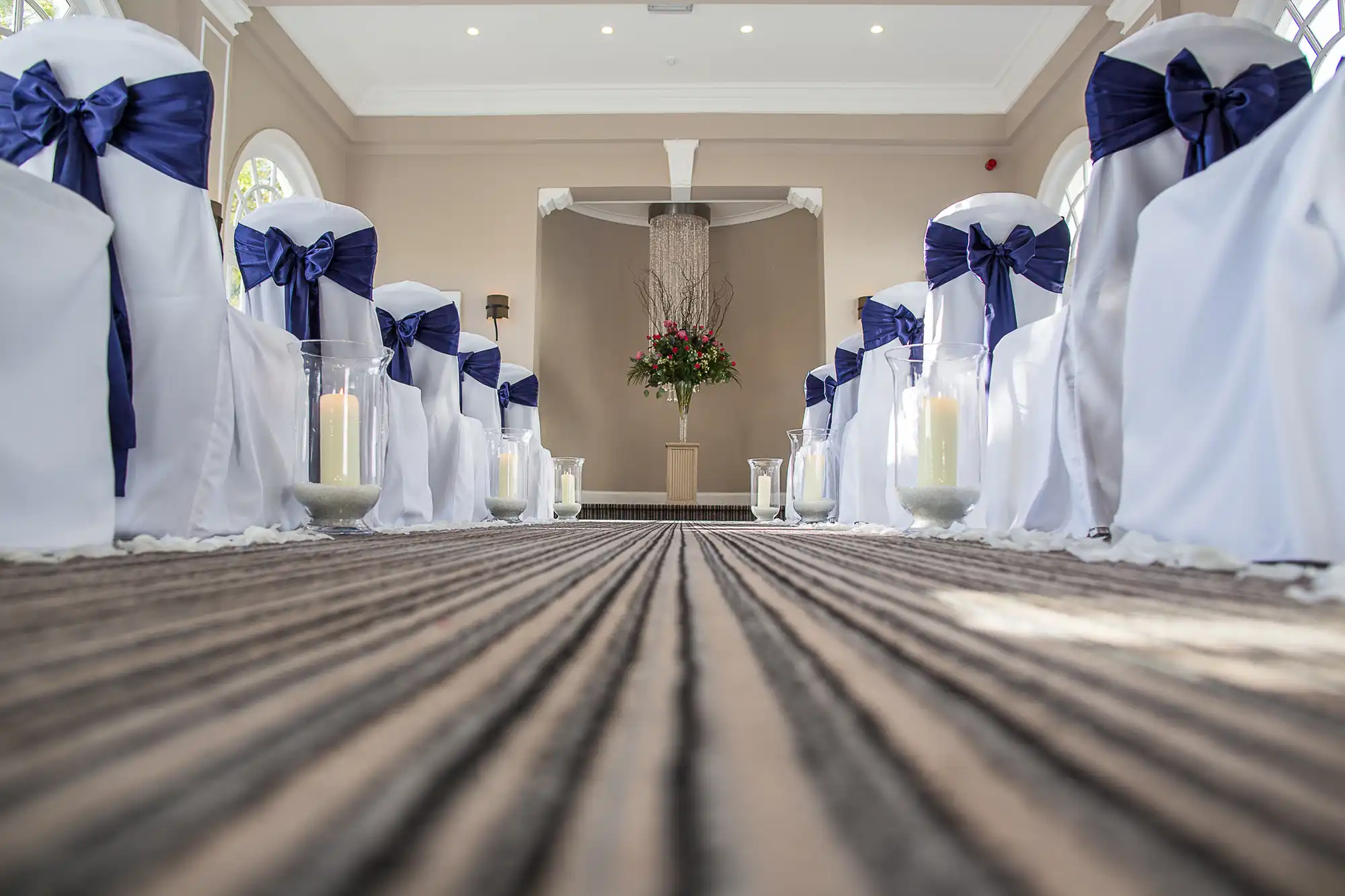 Aisle in a wedding hall decorated with white chairs adorned with large blue bows, and lined with candles on the floor leading to a flower arrangement at the altar.