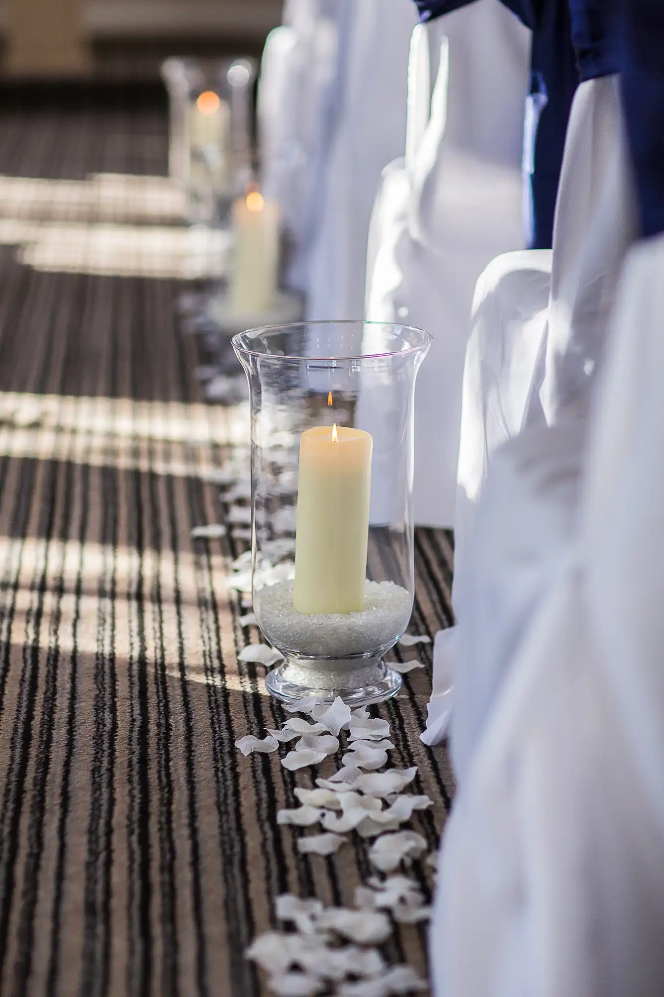 Aisle at a wedding venue decorated with lit candles in glass holders and white petals, alongside chairs with blue sashes.