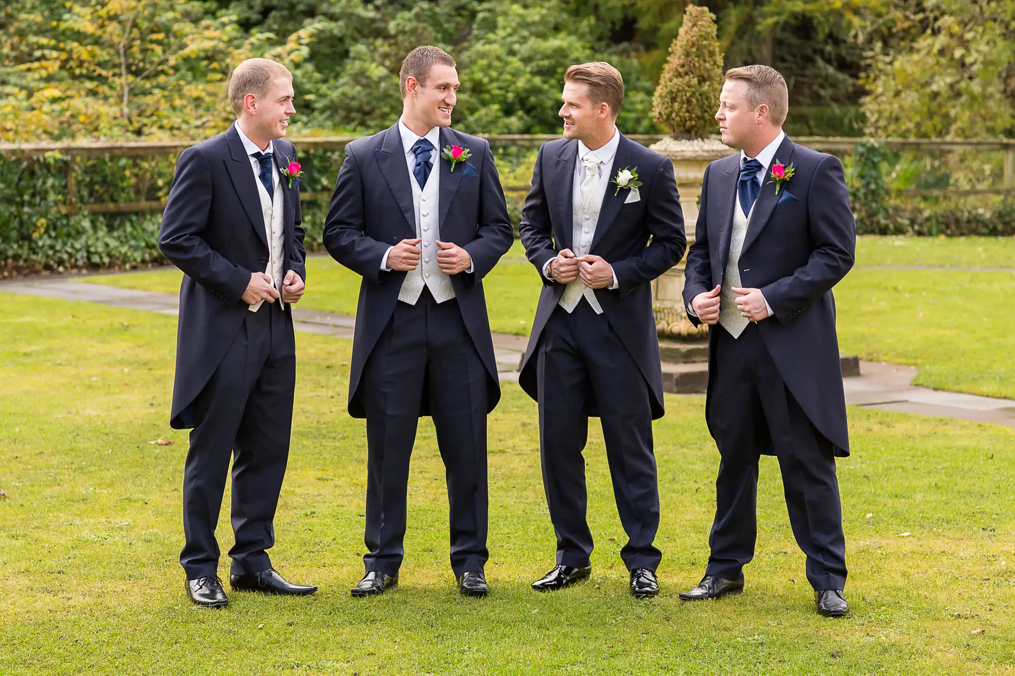 Four men in suits, one wearing a boutonniere, conversing in a garden setting with a gentle smile.