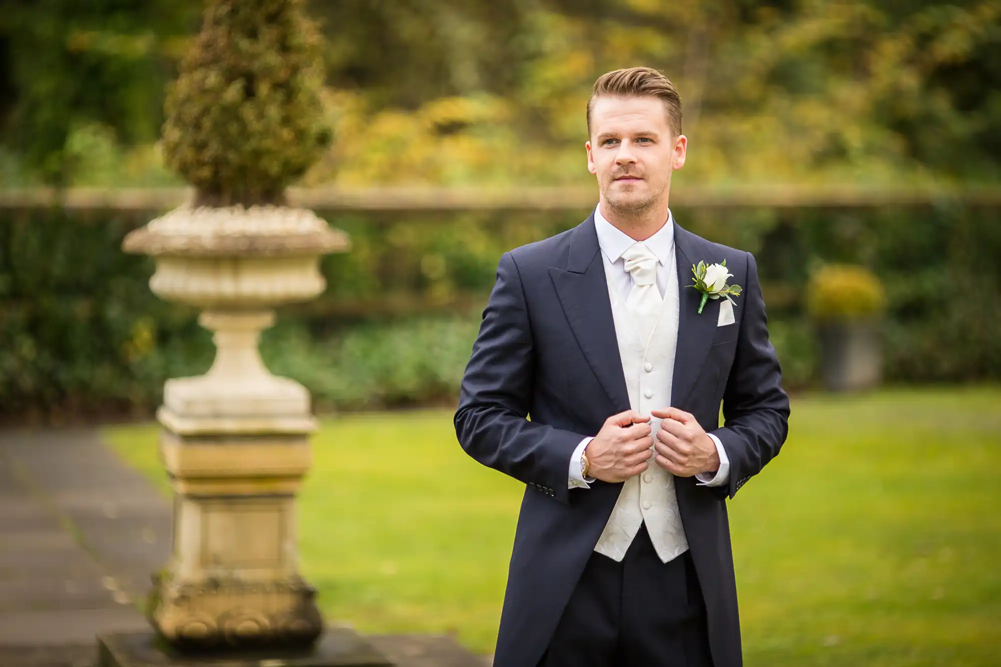 A groom dressed in a formal suit with a boutonniere stands confidently in a garden.