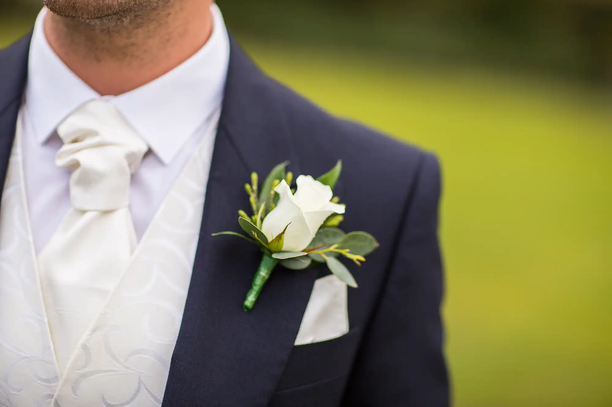 Close-up of a man’s chest in a navy suit with a white rose boutonniere and a silk cravat.