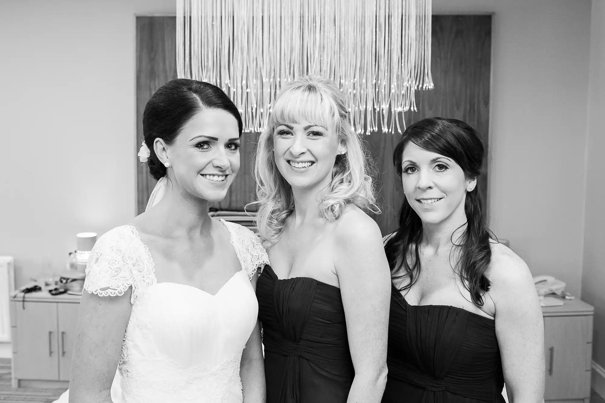 Three women dressed for a special occasion, posing together smiling; one in a white bridal gown, two in black dresses, in a room with a modern chandelier.