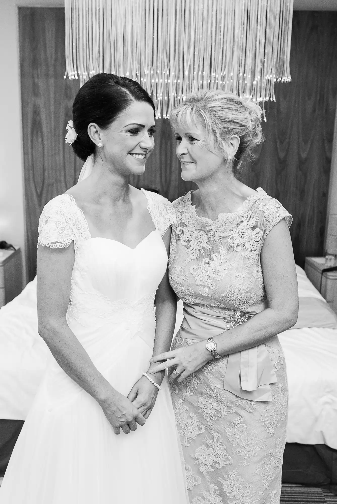 A black and white photo of a bride in a lace dress smiling at an older woman in a lace dress, possibly her mother, as they hold hands.