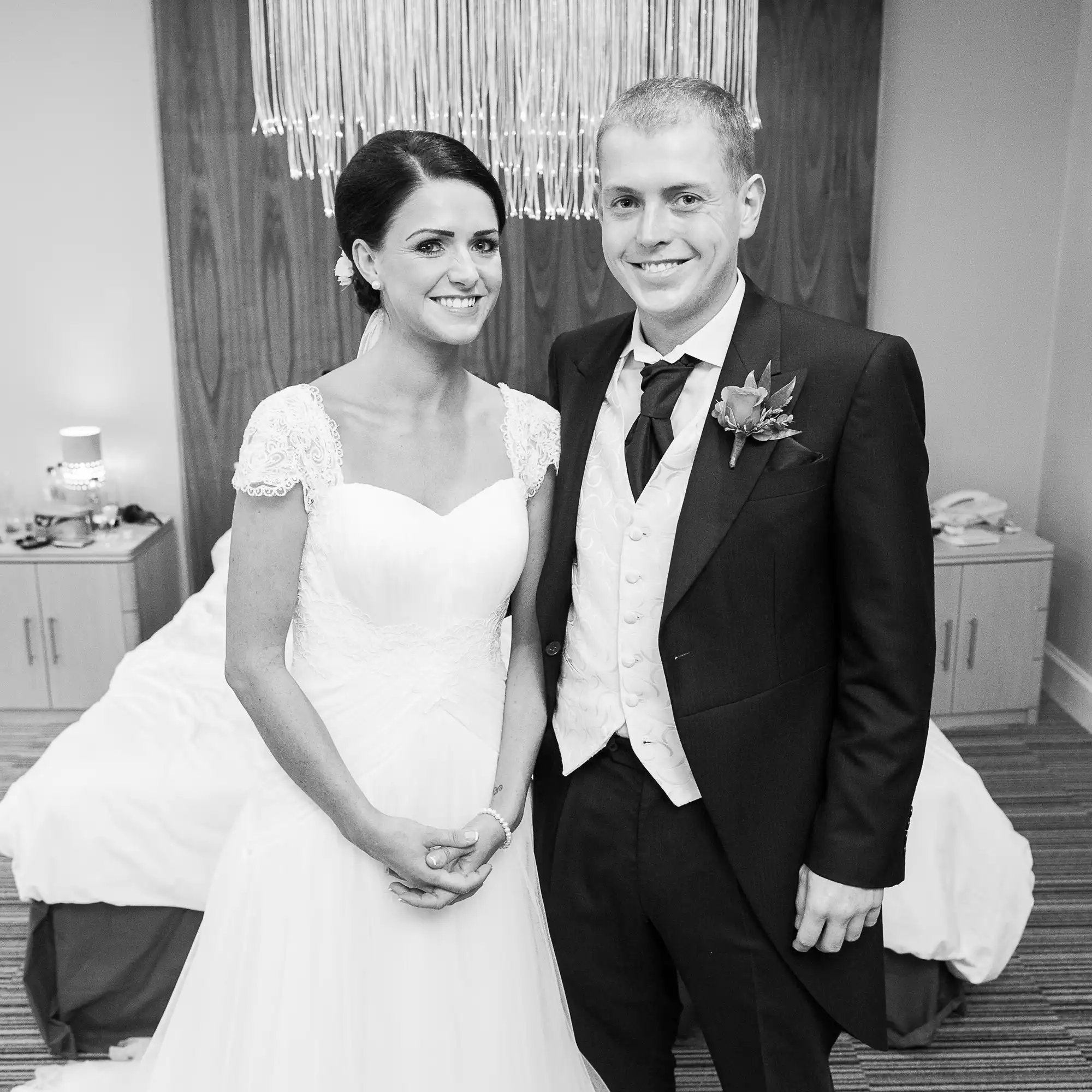 A smiling bride in a lace dress and a groom in a suit holding hands at their wedding, in a black and white photo.