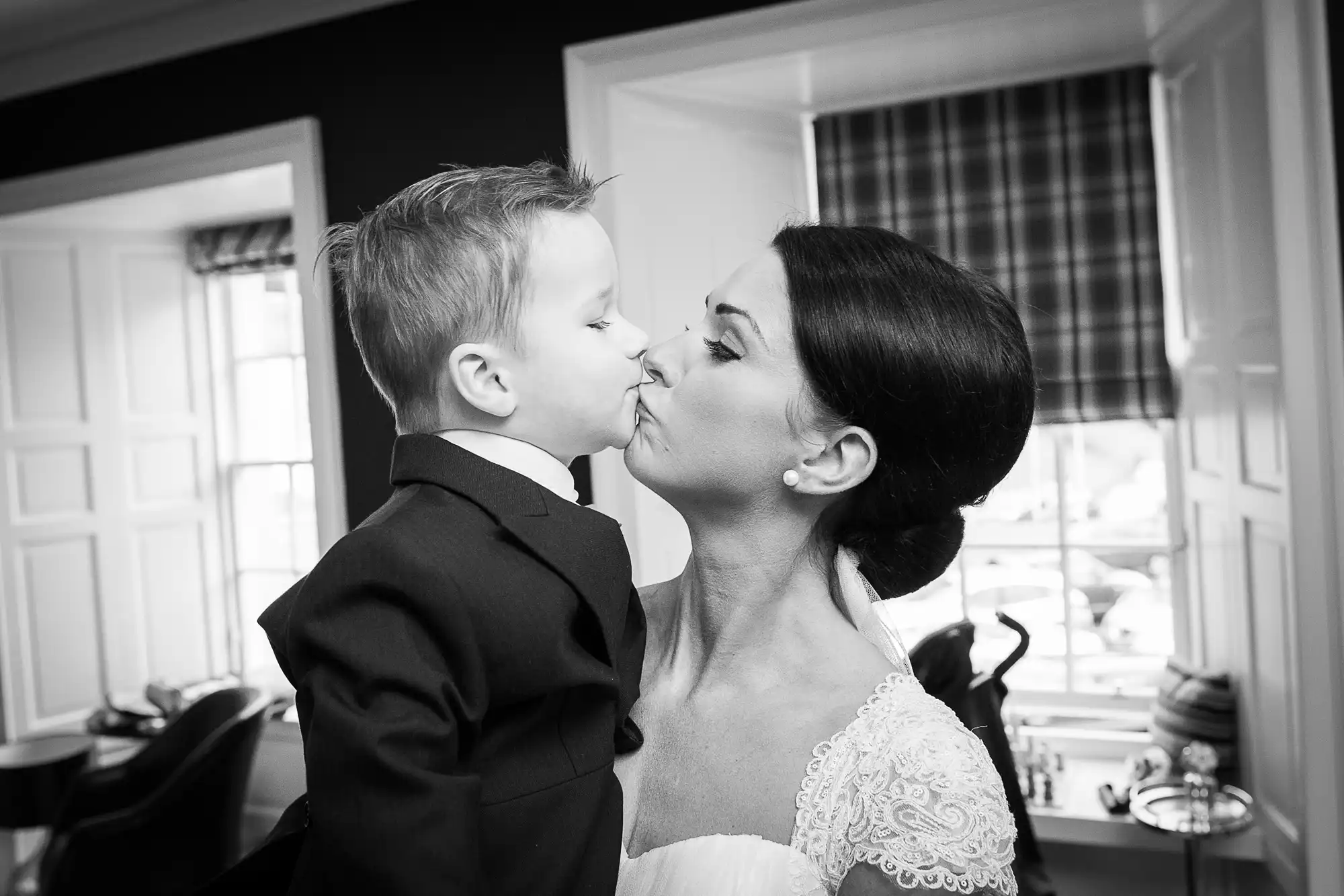 A black and white photo of a young boy in a suit kissing a woman in a lace wedding dress on the cheek.