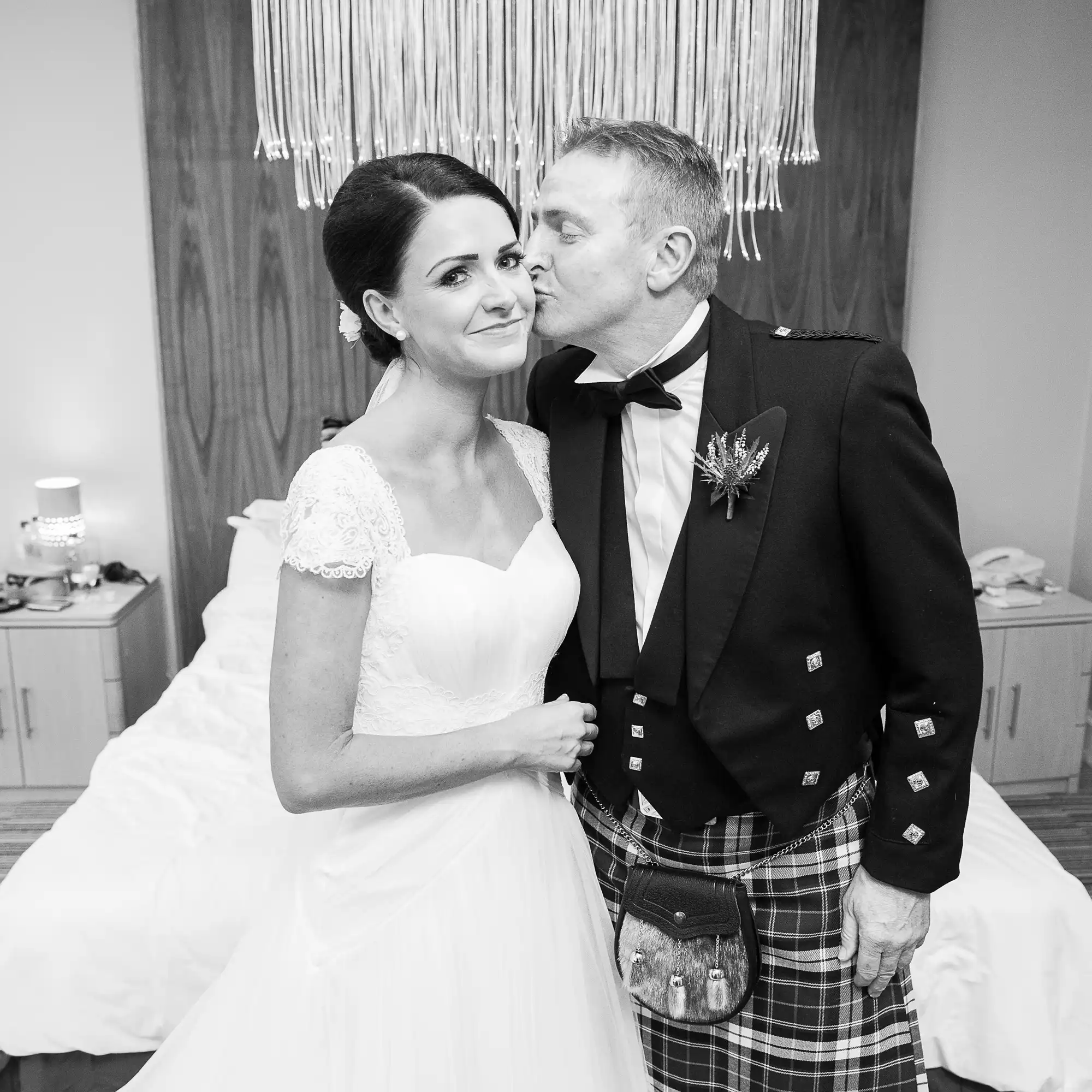 A man in a kilt kissing a bride on the cheek in a black and white wedding photo.