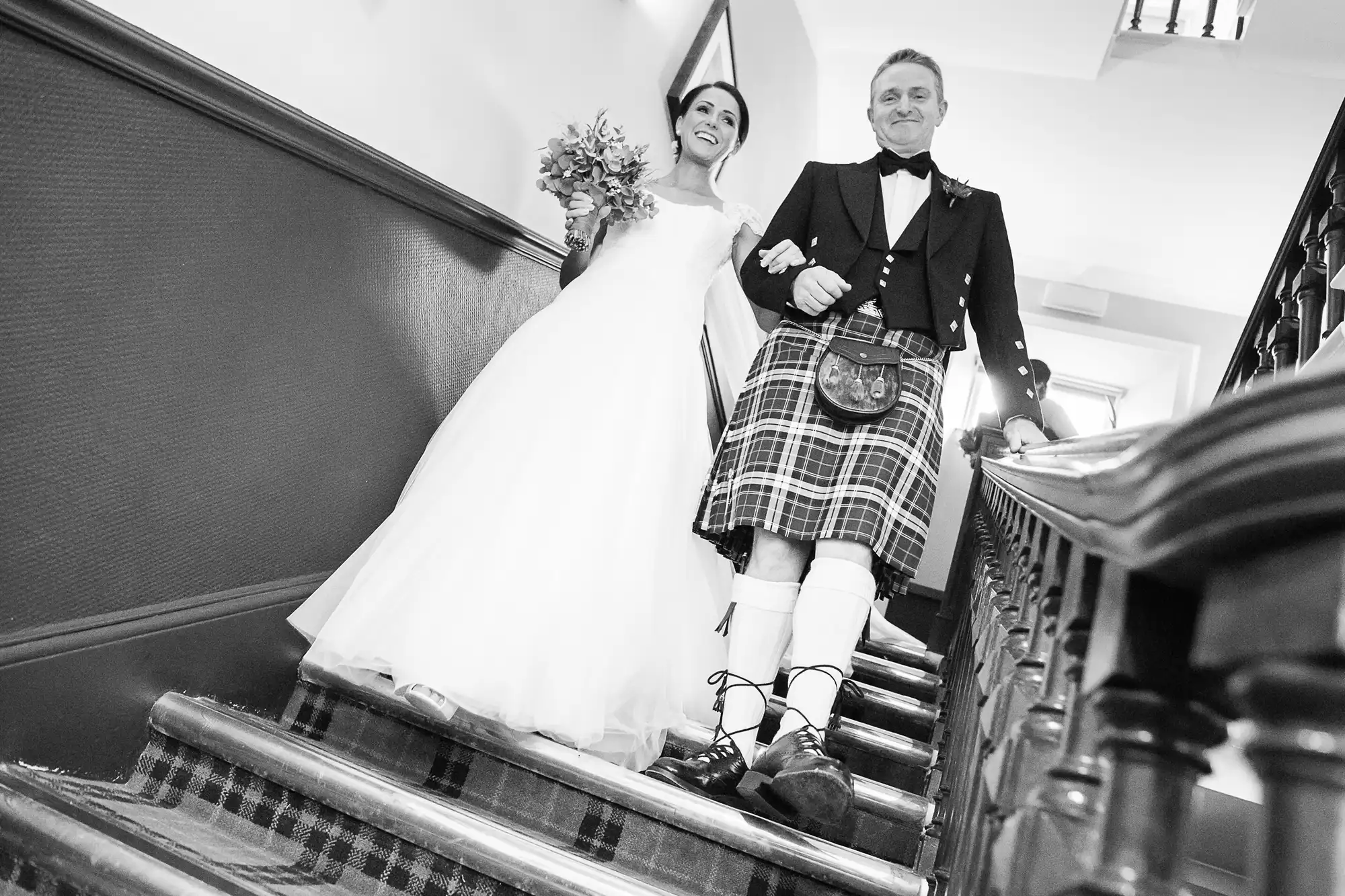 A bride in a white dress and a groom in a traditional kilt descend a staircase, both smiling broadly.