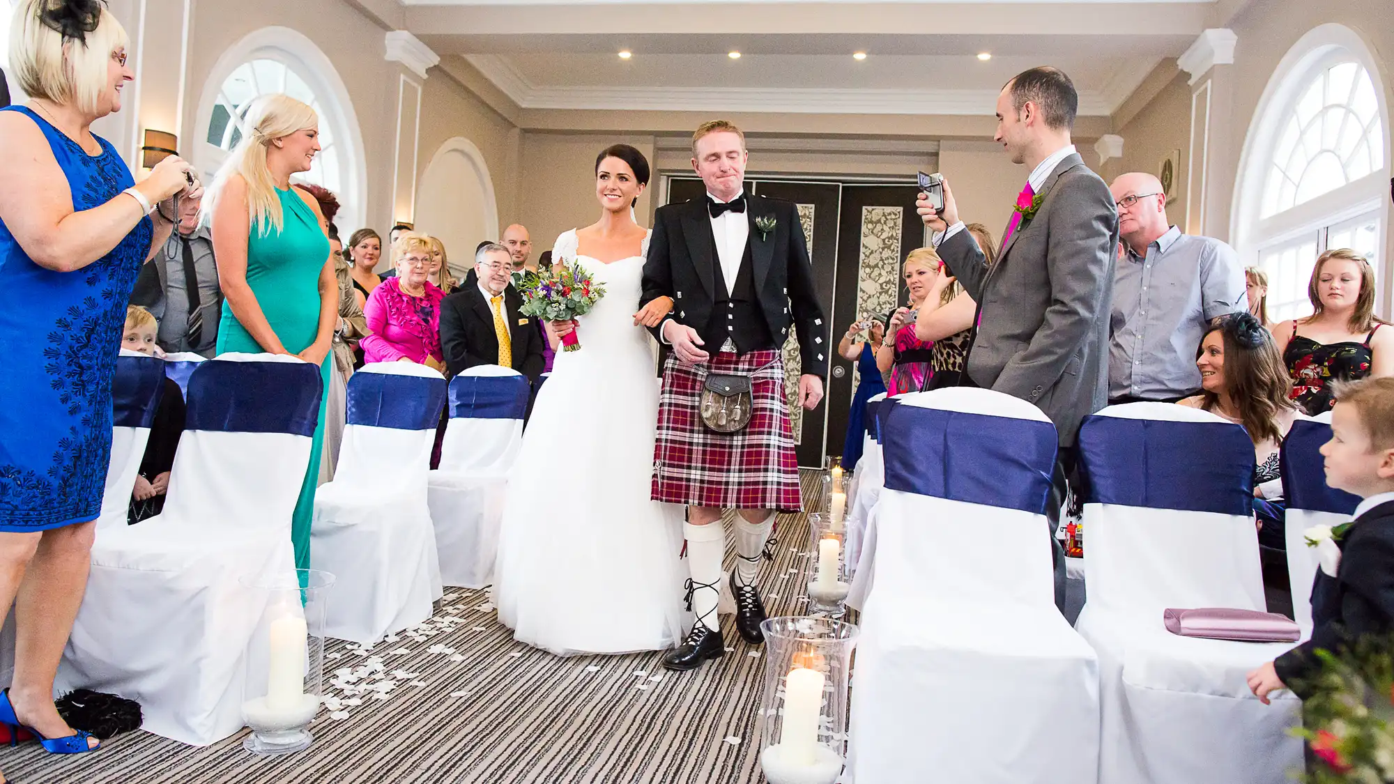 A bride and groom walking down the aisle, the groom wearing a kilt, with guests watching and taking photos in a decorated wedding venue.