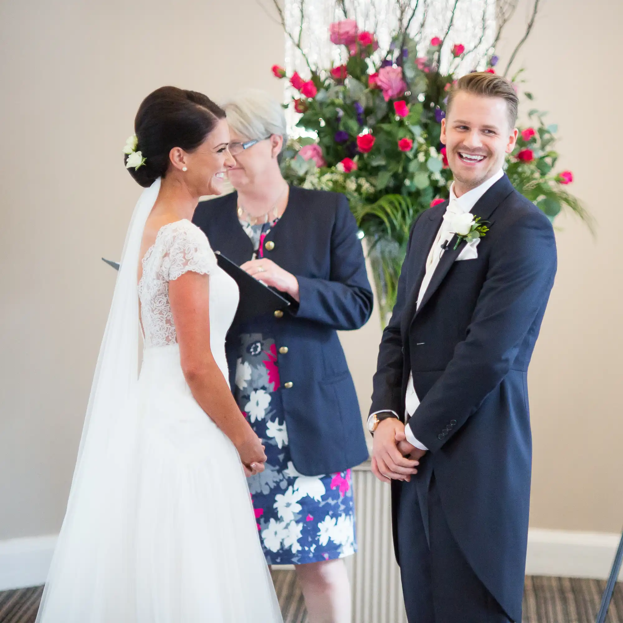 A bride and groom smiling at each other during their wedding ceremony, with an officiant in the background.