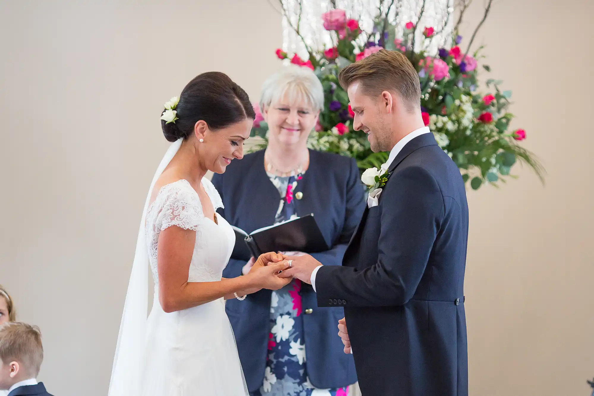 Bride and groom exchanging rings during a wedding ceremony, with an officiant smiling in the background.