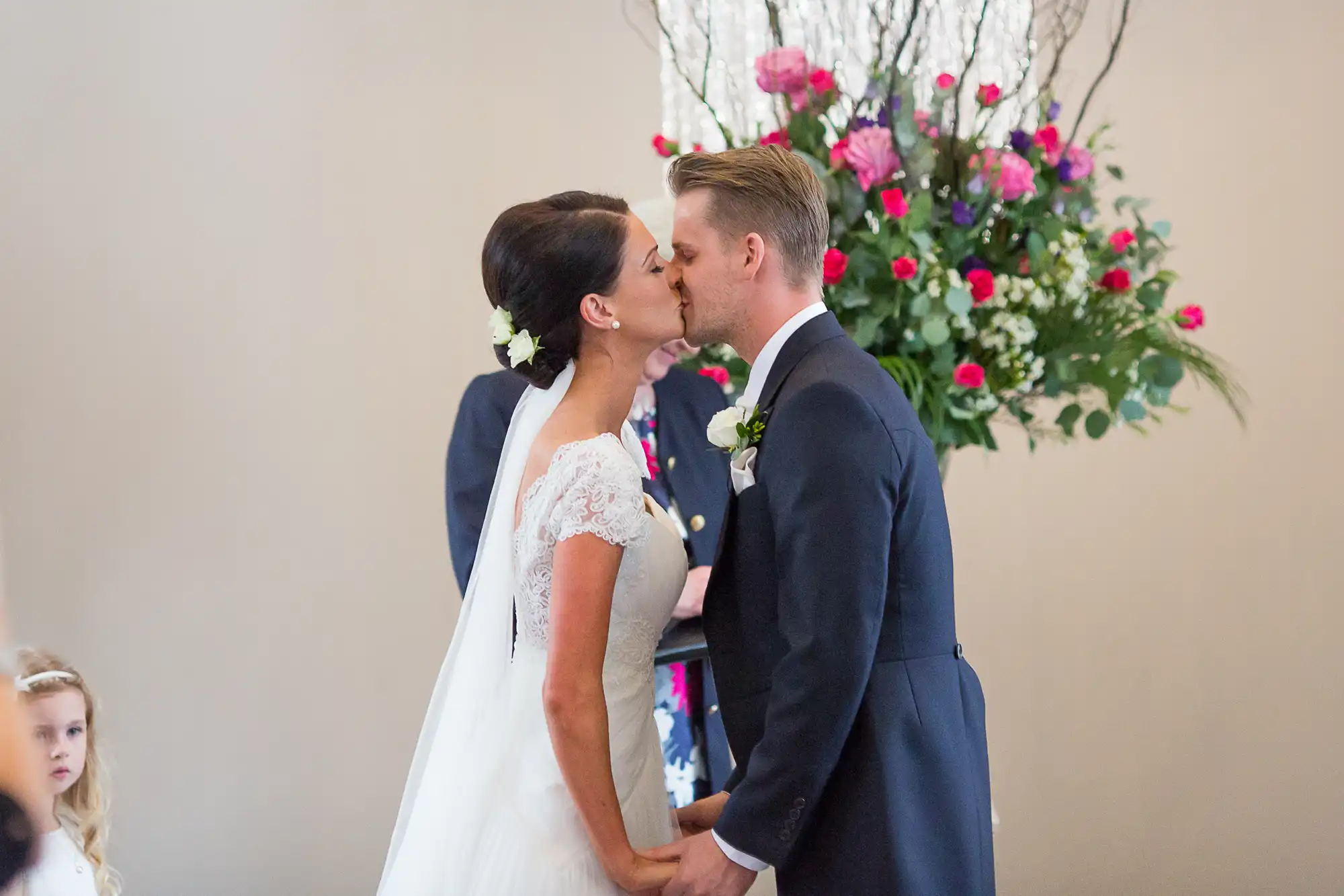A bride and groom kissing at the altar, with vibrant flowers in the background and a young bridesmaid watching.