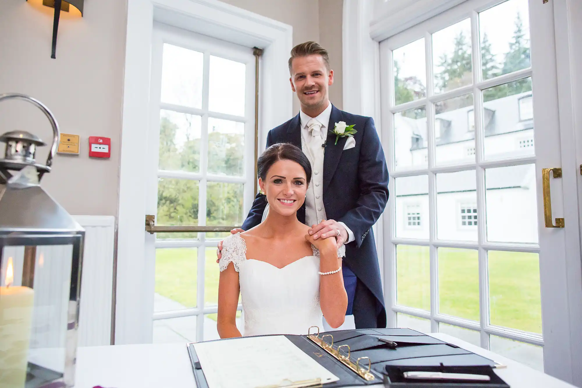 A bride and groom smiling at the camera, standing behind a signing table with documents in a bright room with windows.
