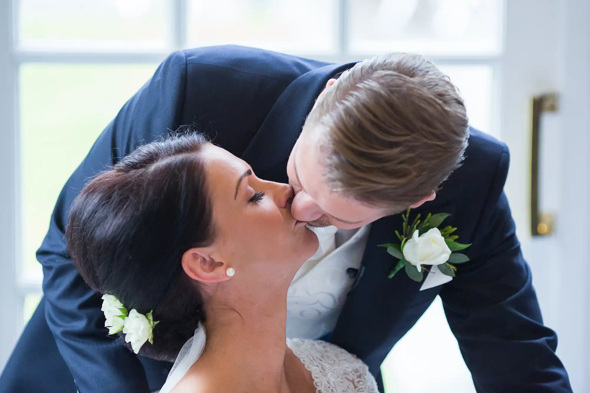 A groom bending down to kiss his bride on the forehead in a sunlit room.