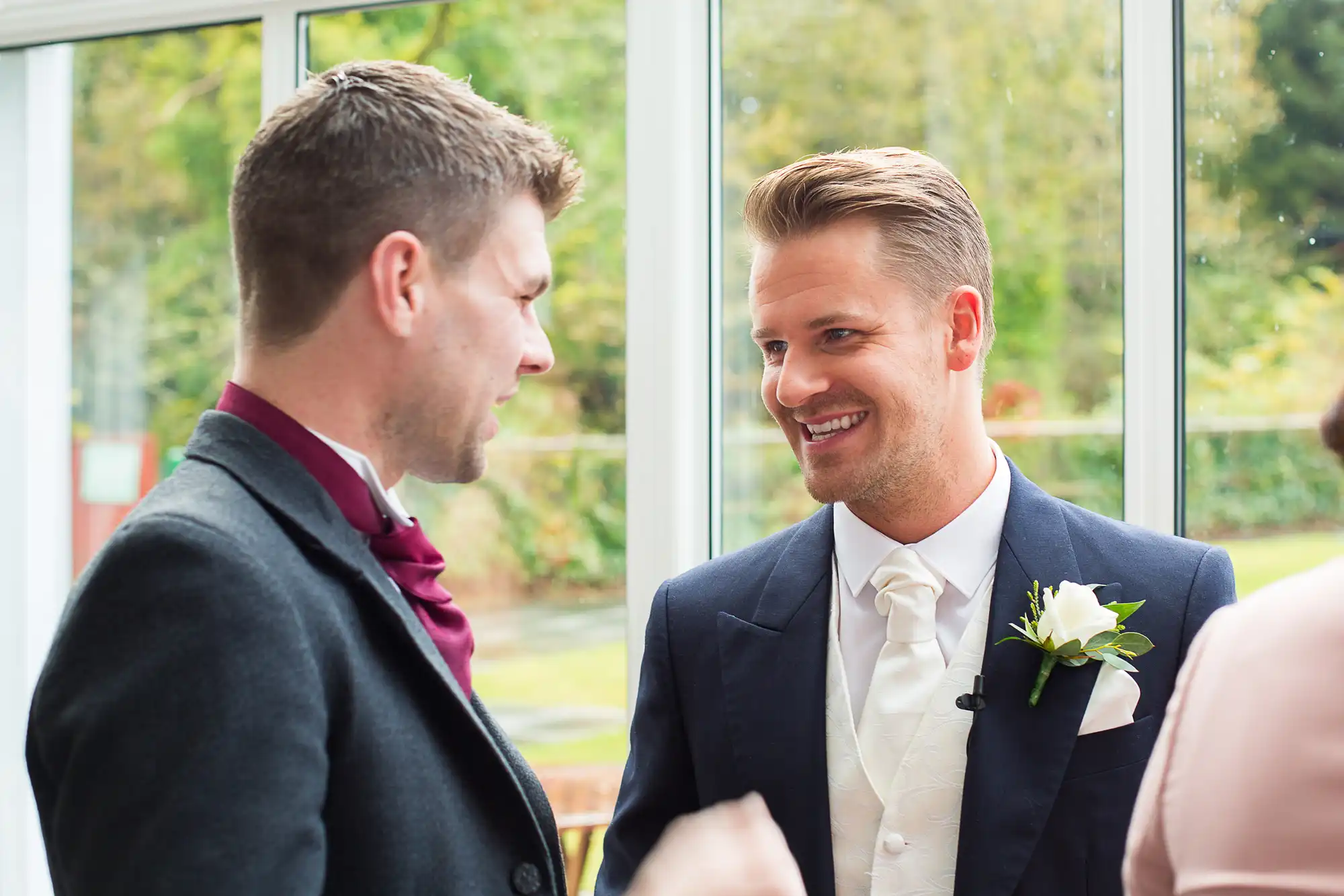 Two men in formal attire smiling and conversing indoors near a window during a daytime event. one wears a boutonniere.