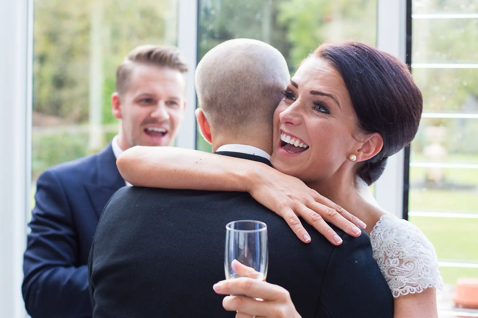 A woman with a glass of wine embracing a bald man at a gathering, smiling joyfully, while a young man in the background watches, also smiling.