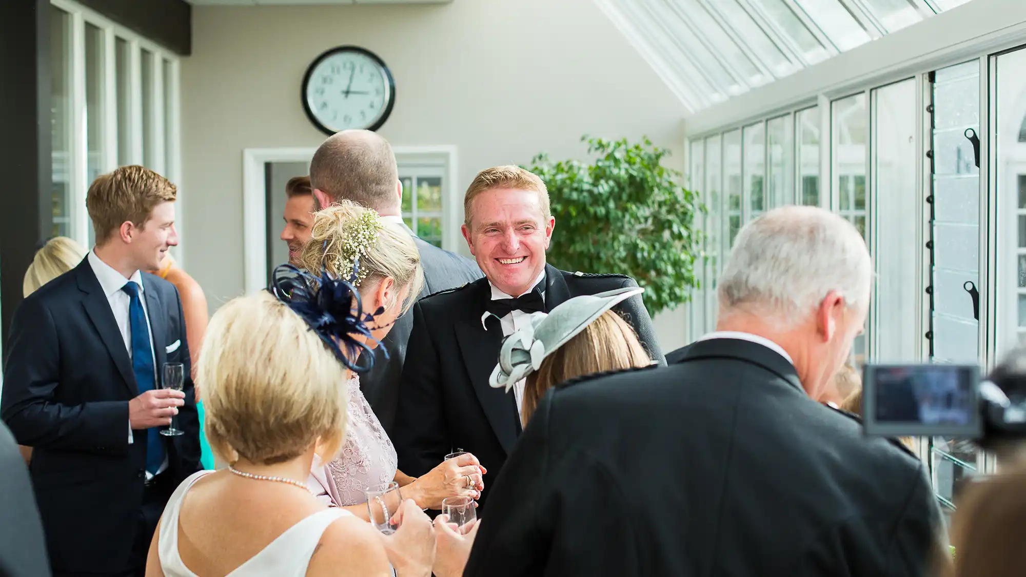 A man smiles at a wedding reception filled with guests, including some wearing fascinators, in a well-lit conservatory-style room.