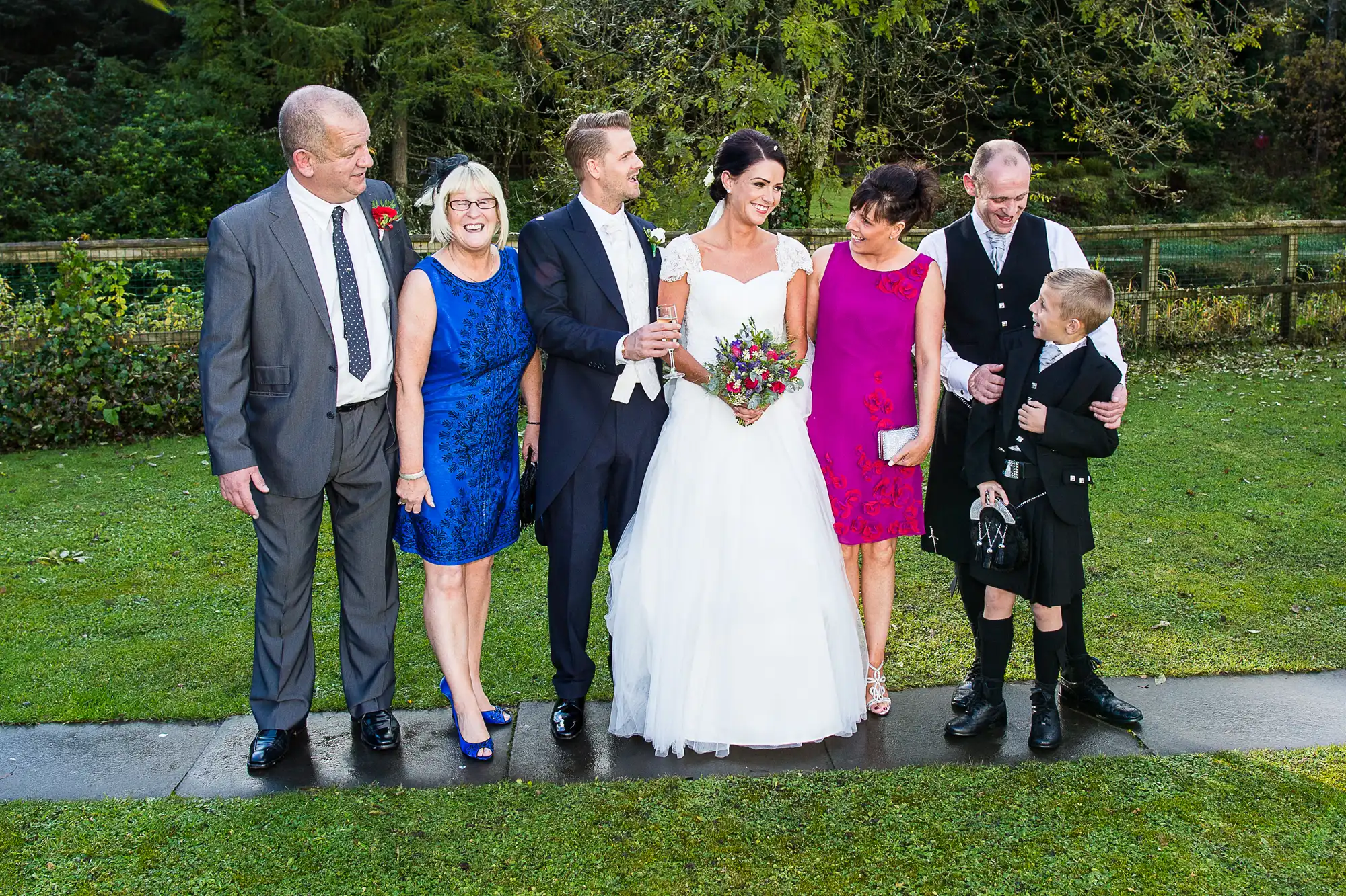 A wedding group photo featuring a bride, groom, and five guests, including two children, smiling outdoors on a sunny day.