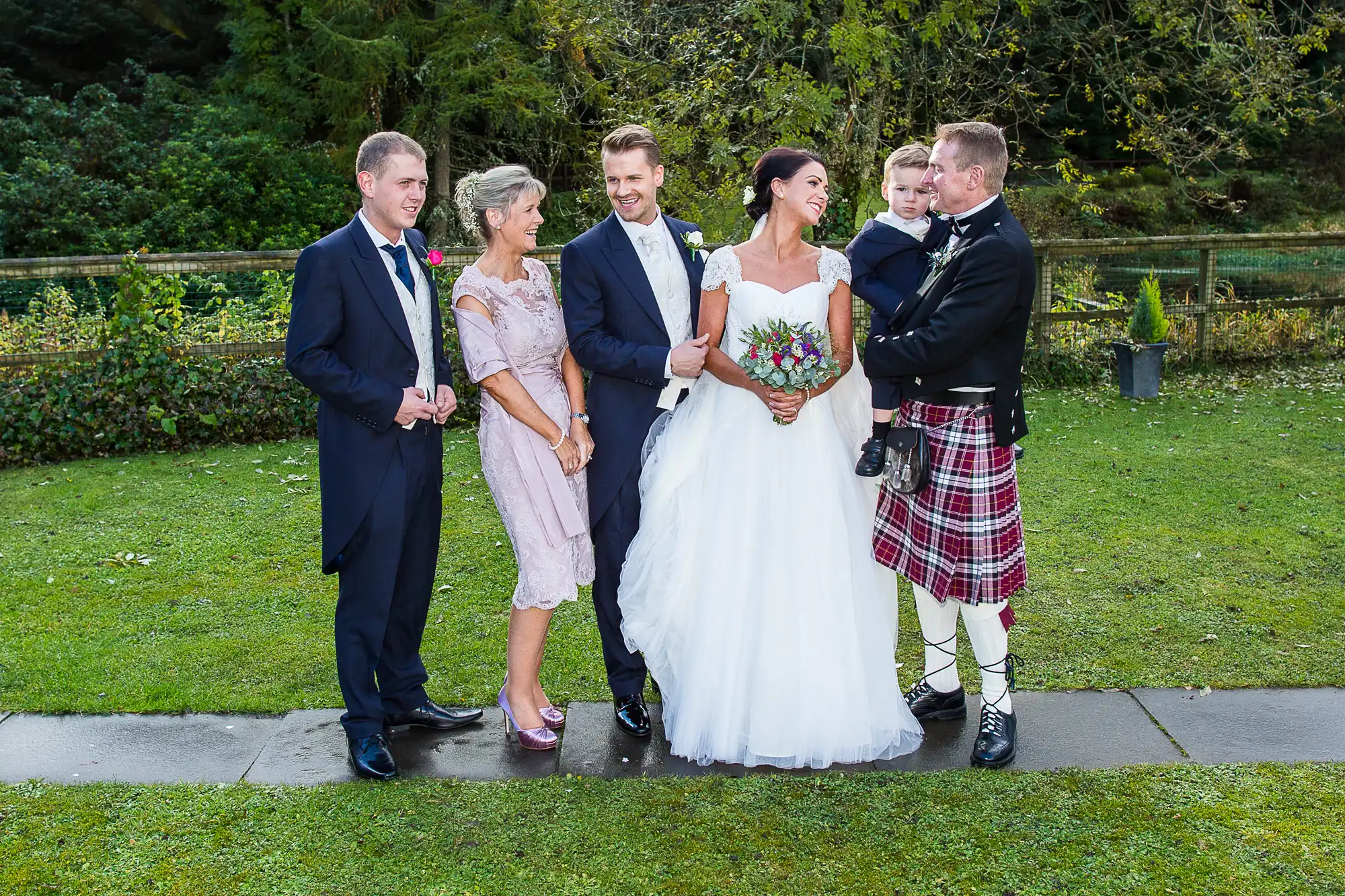 A wedding photo featuring three men and two women, including a bride in a white gown and a man in a kilt, standing on grass with trees in the background.