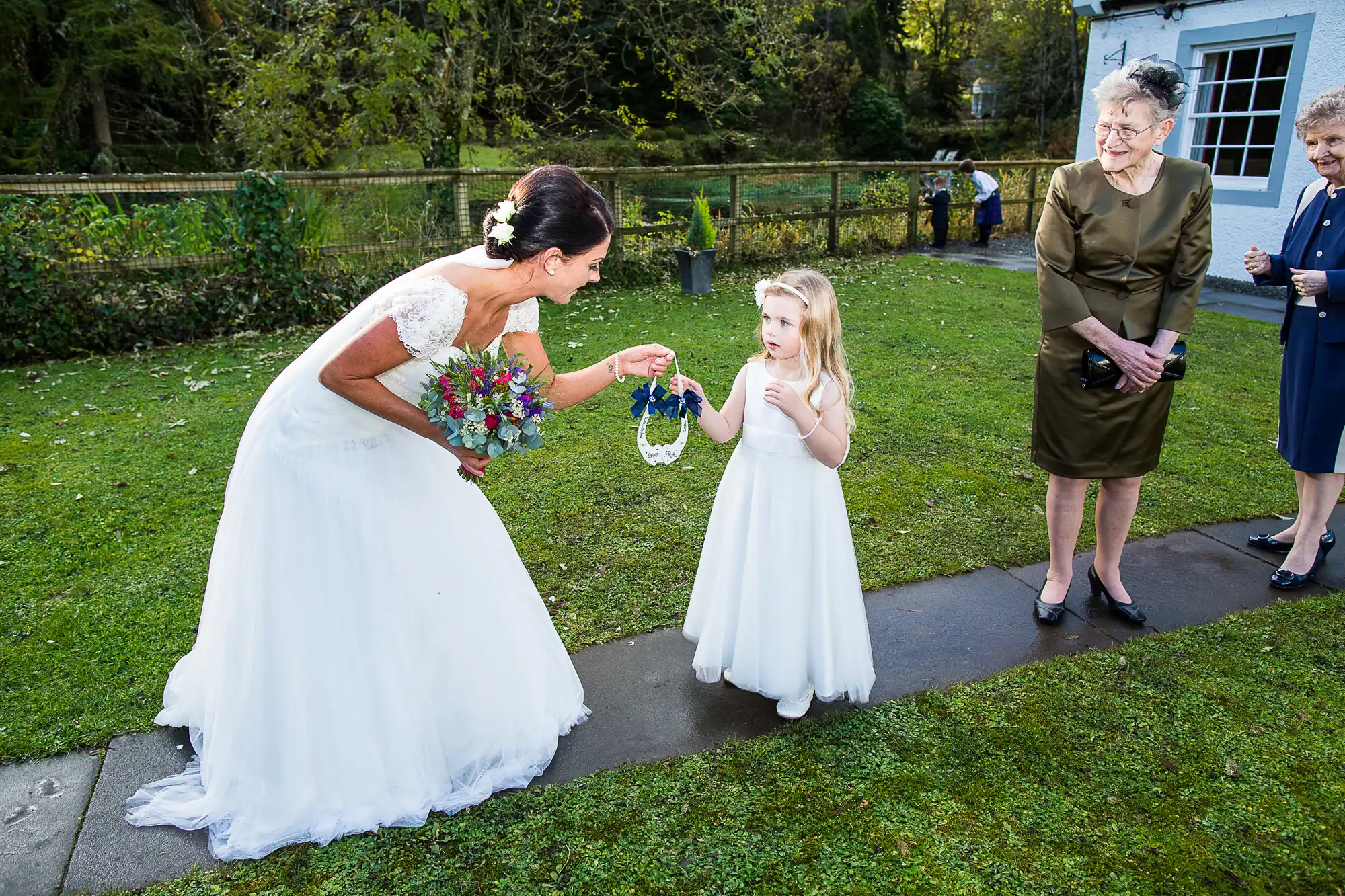 A bride in a white gown leans down to talk to a young flower girl in a white dress, with two older women looking on, in an outdoor setting.