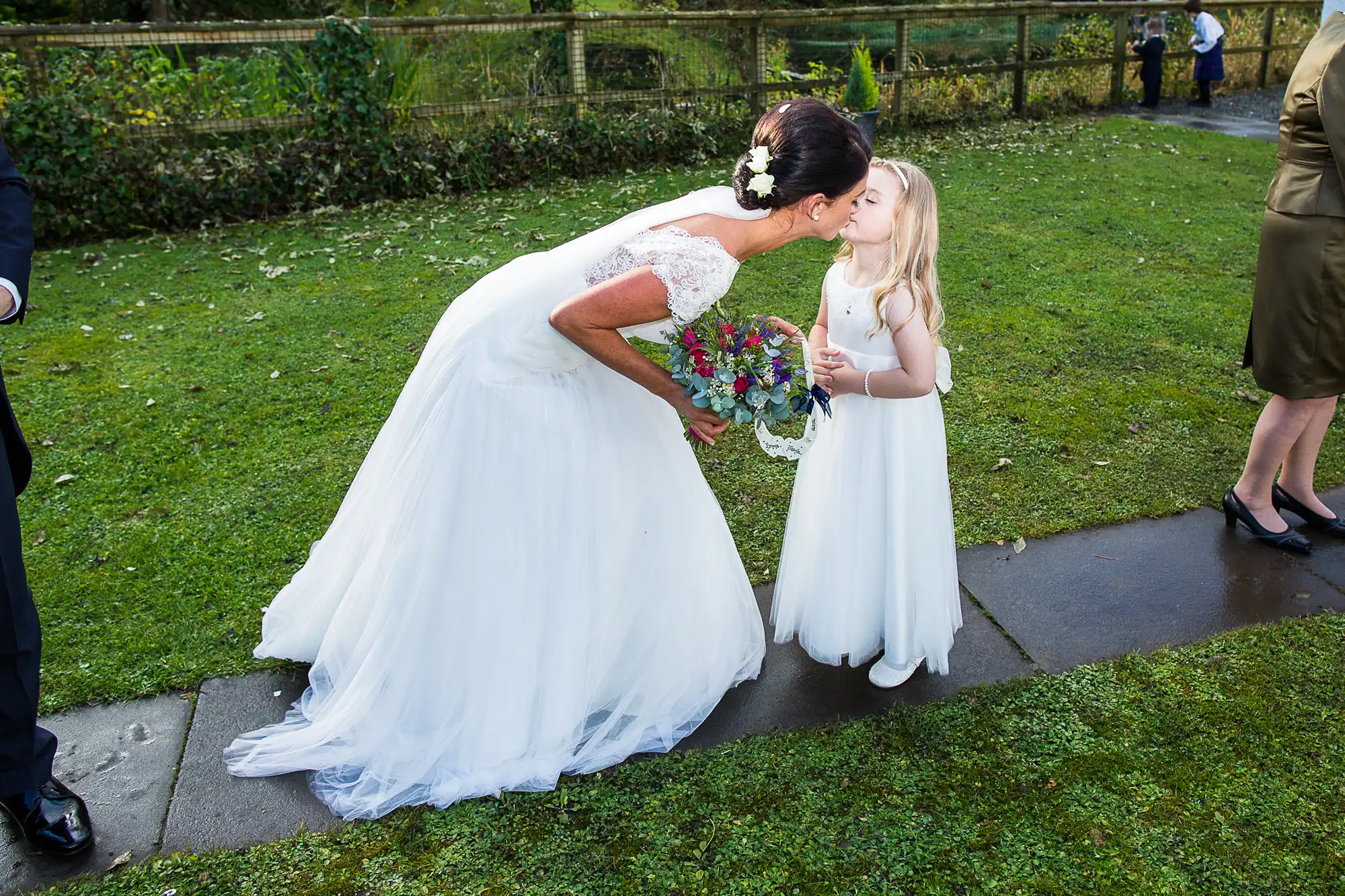 A bride in a white gown kisses a young girl in a white dress holding a bouquet, outdoors on grass by a fence.