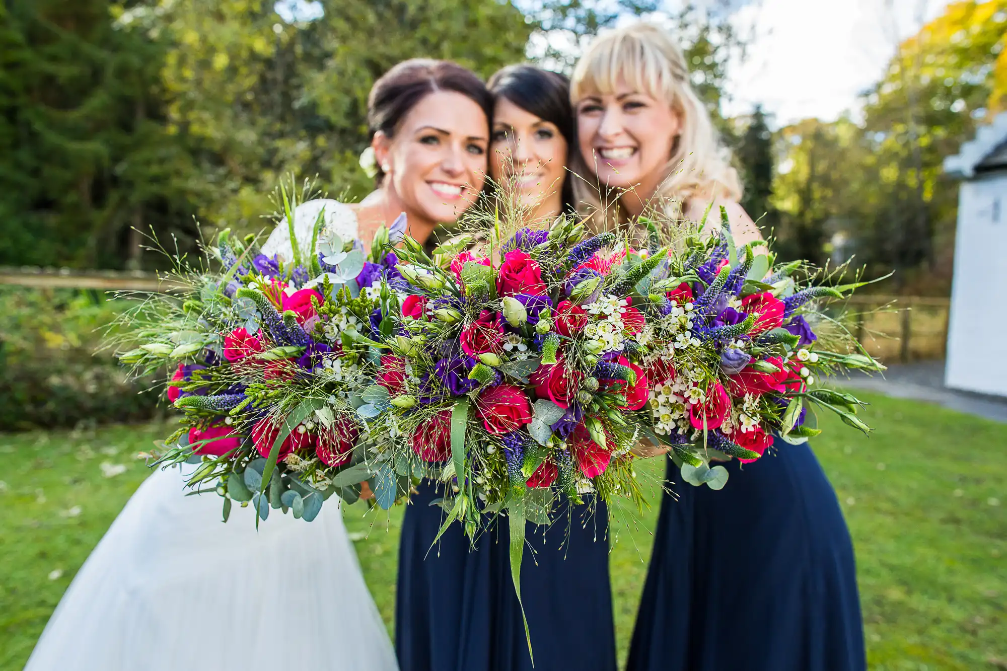 Three smiling women at a wedding, one in a white dress holding a large, colorful floral bouquet, flanked by two in blue dresses.