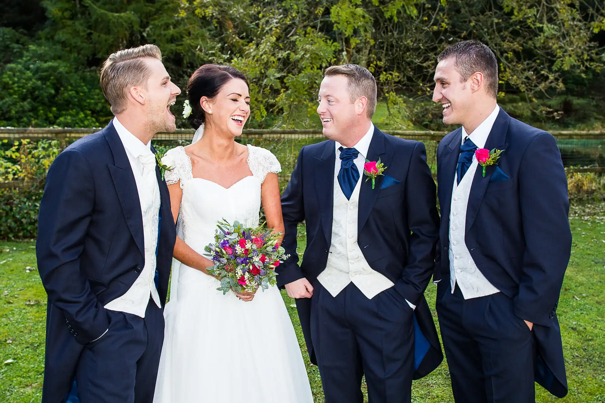 A bride and three men in suits laughing together outdoors, with the bride holding a colorful bouquet.