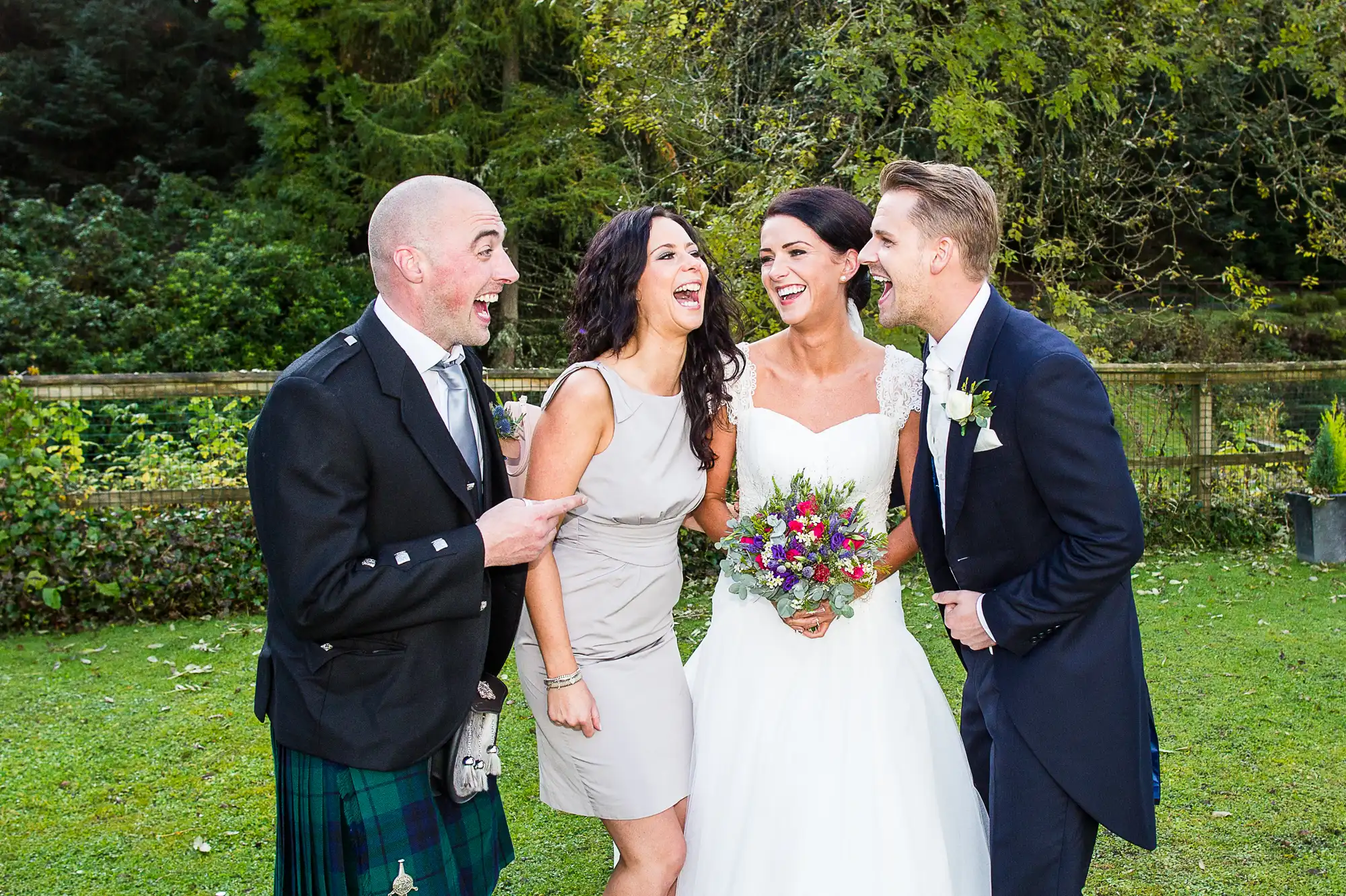 A joyful wedding group outdoors, featuring a laughing couple in traditional and modern wedding attire, joined by two happy friends.