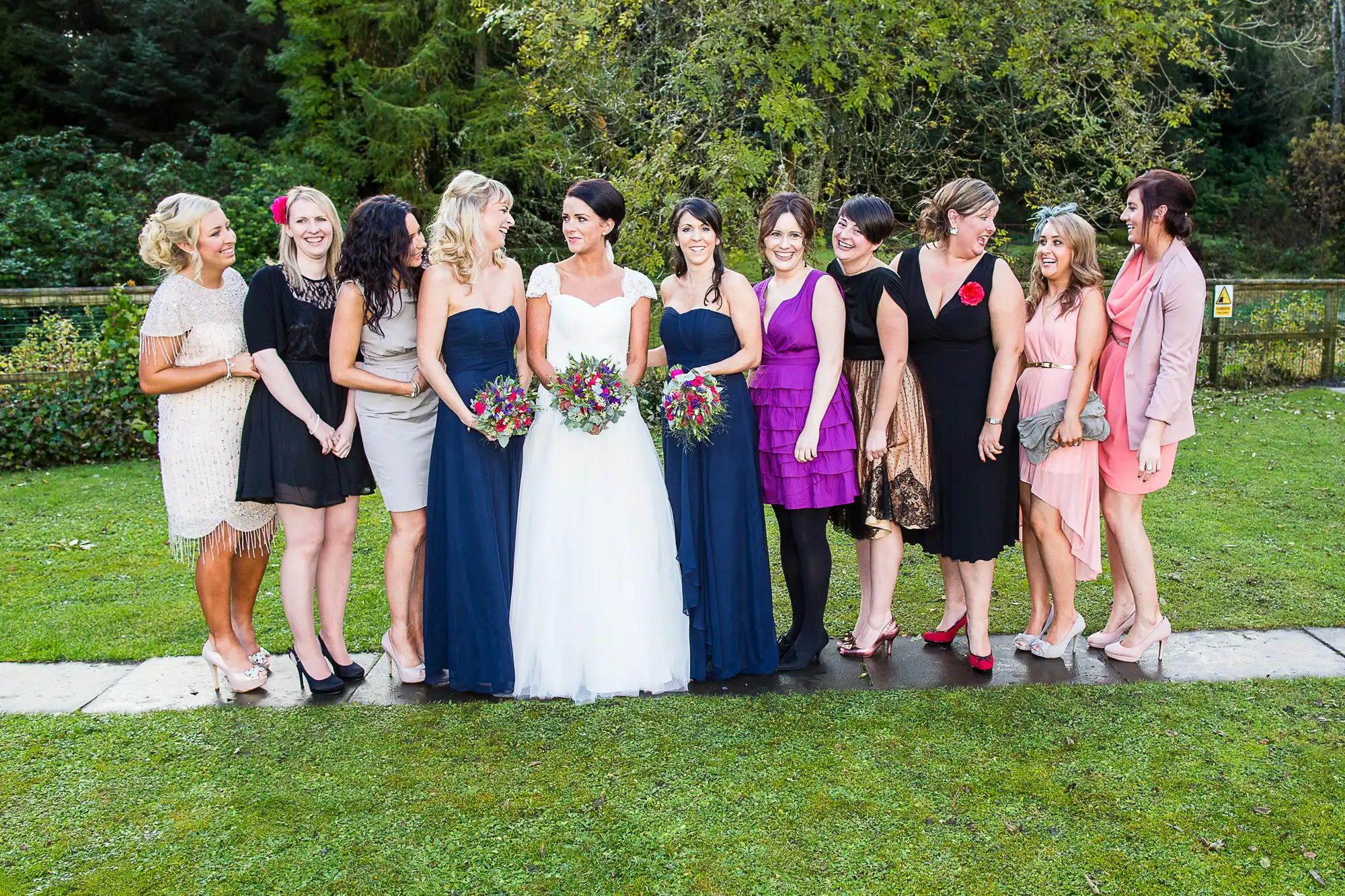 A bride in a white dress stands with nine women in various colored dresses in a garden, all smiling.