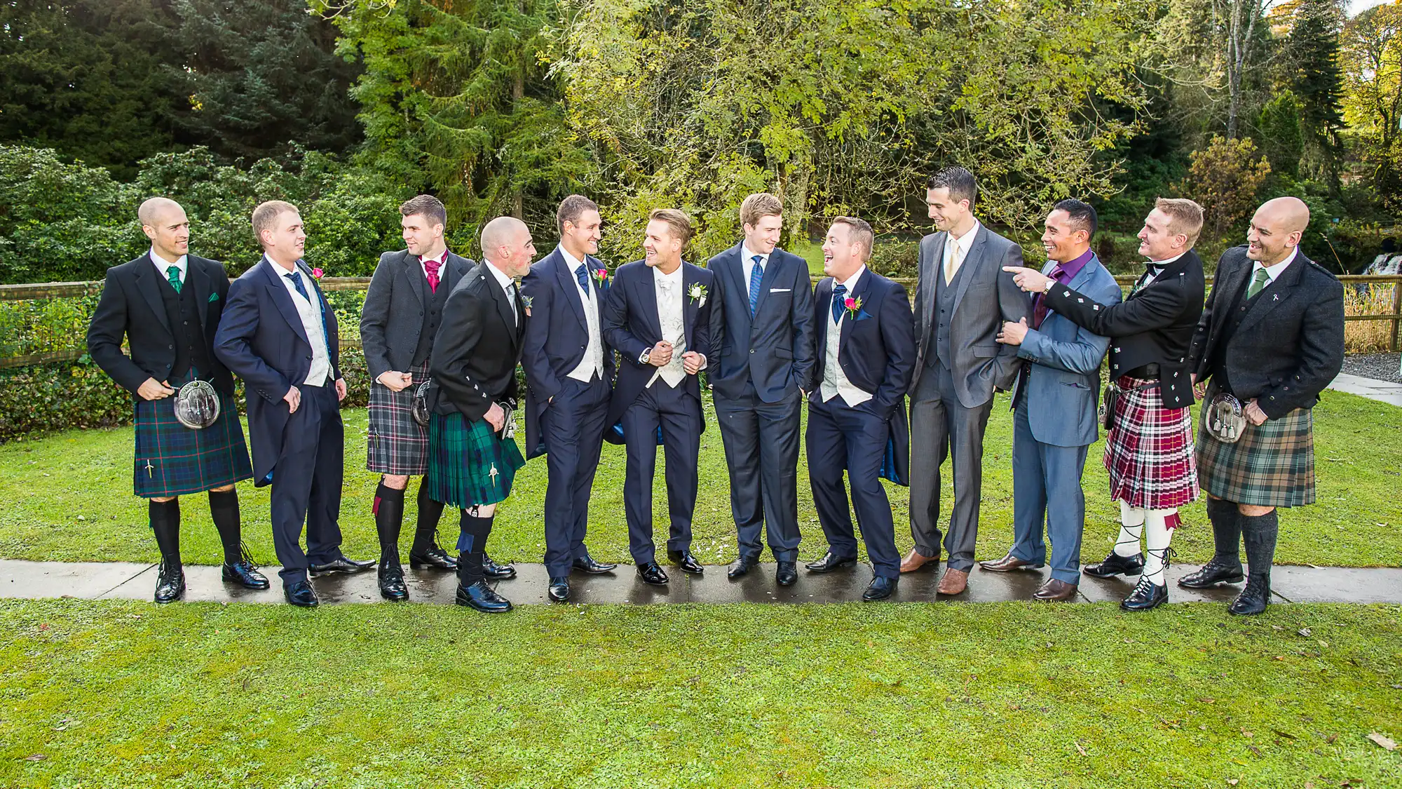 Group of men in suits and kilts smiling and interacting at an outdoor event, surrounded by greenery.