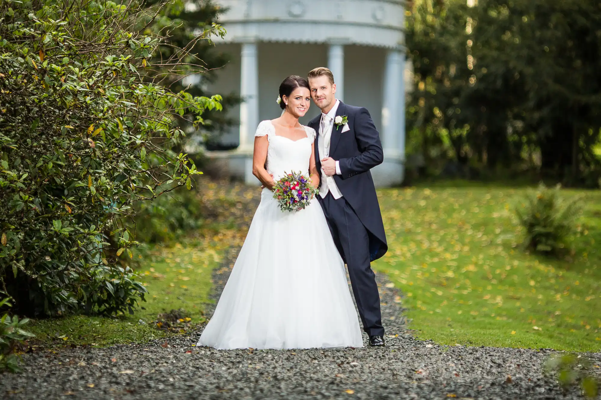 A bride and groom smiling and posing in a garden pathway, with the bride holding a colorful bouquet.