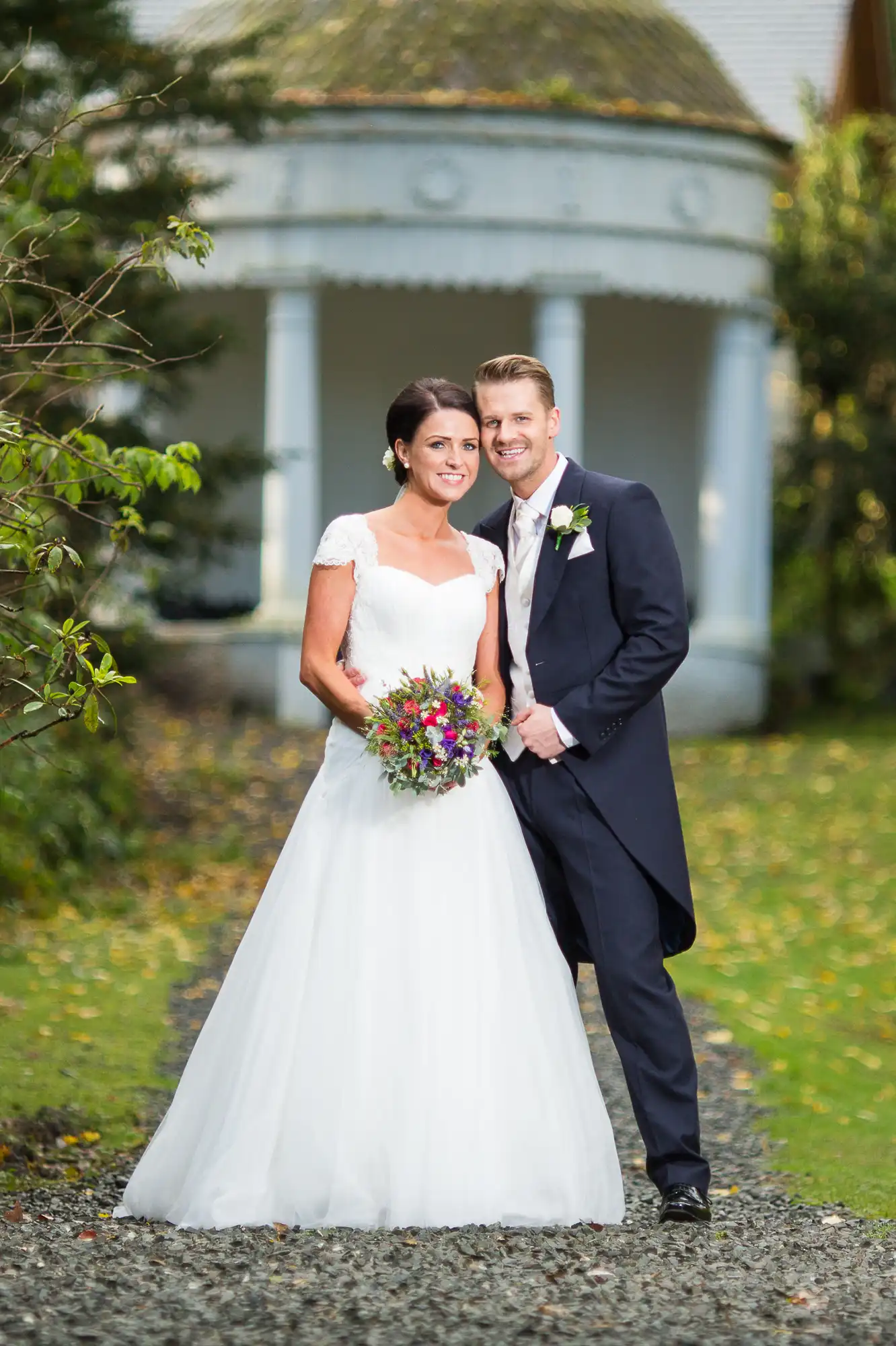 A bride and groom smiling and posing in front of a gazebo, the bride in a white wedding dress holding a colorful bouquet, and the groom in a dark suit with a boutonniere.