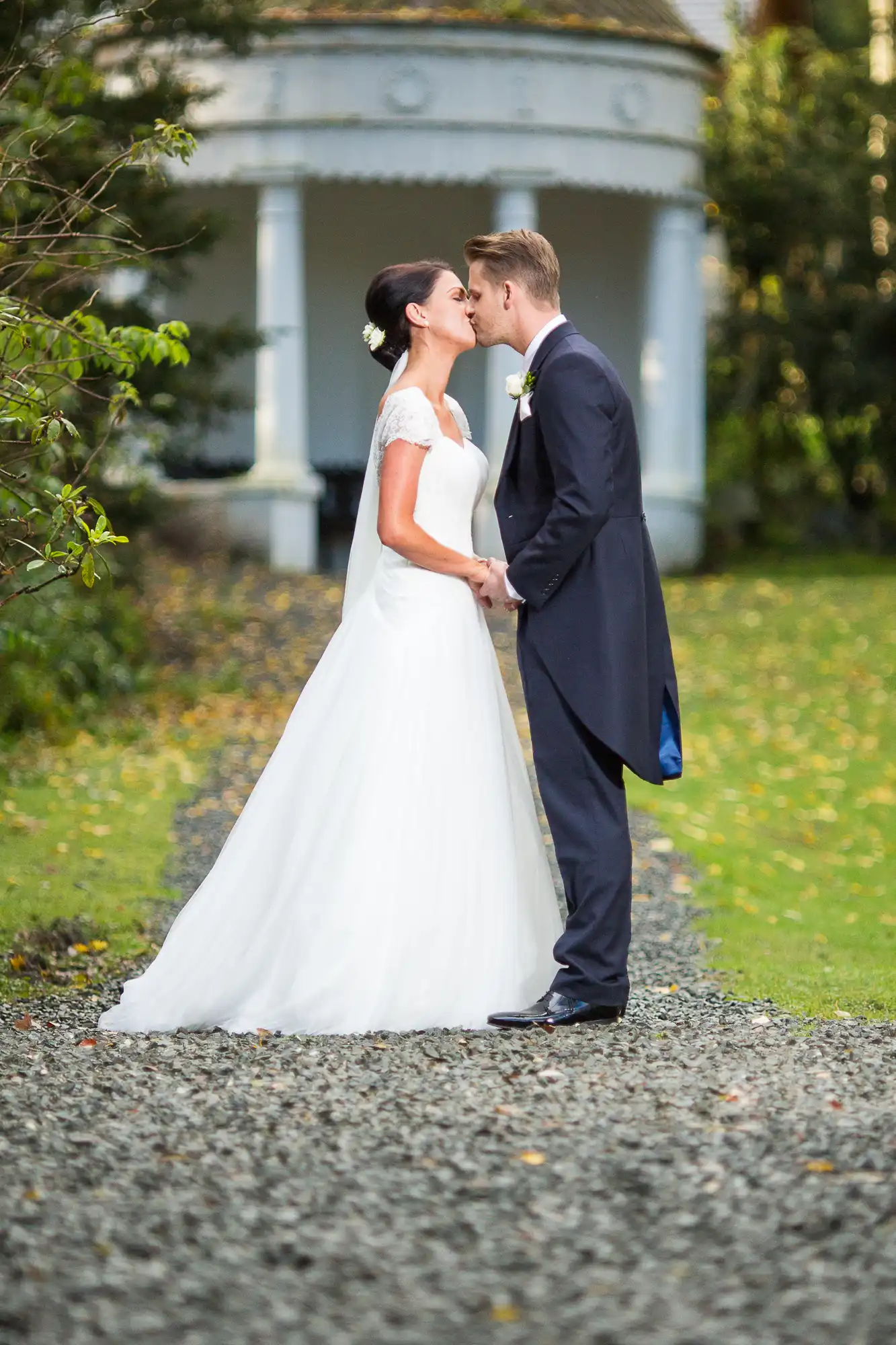 A bride and groom kissing outdoors, with the bride in a white dress and the groom in a navy suit, standing in front of a white gazebo surrounded by trees.