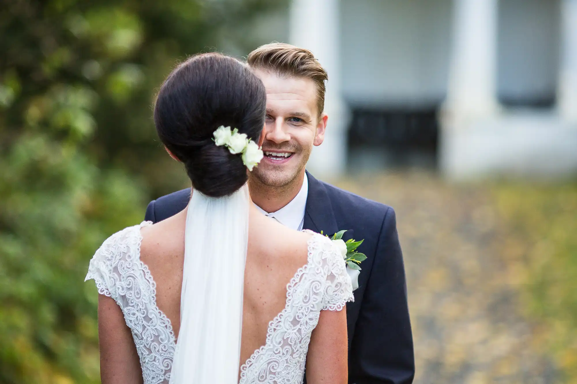 A groom smiling at the camera over the shoulder of a bride with her back facing us, featuring a lace wedding dress and white floral hair accessory.