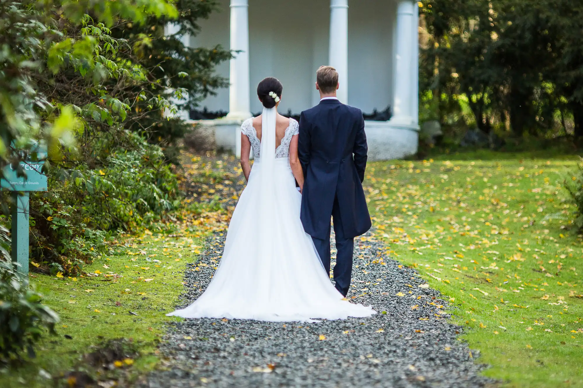 A bride and groom walking hand in hand toward a white gazebo in a garden, viewed from behind.