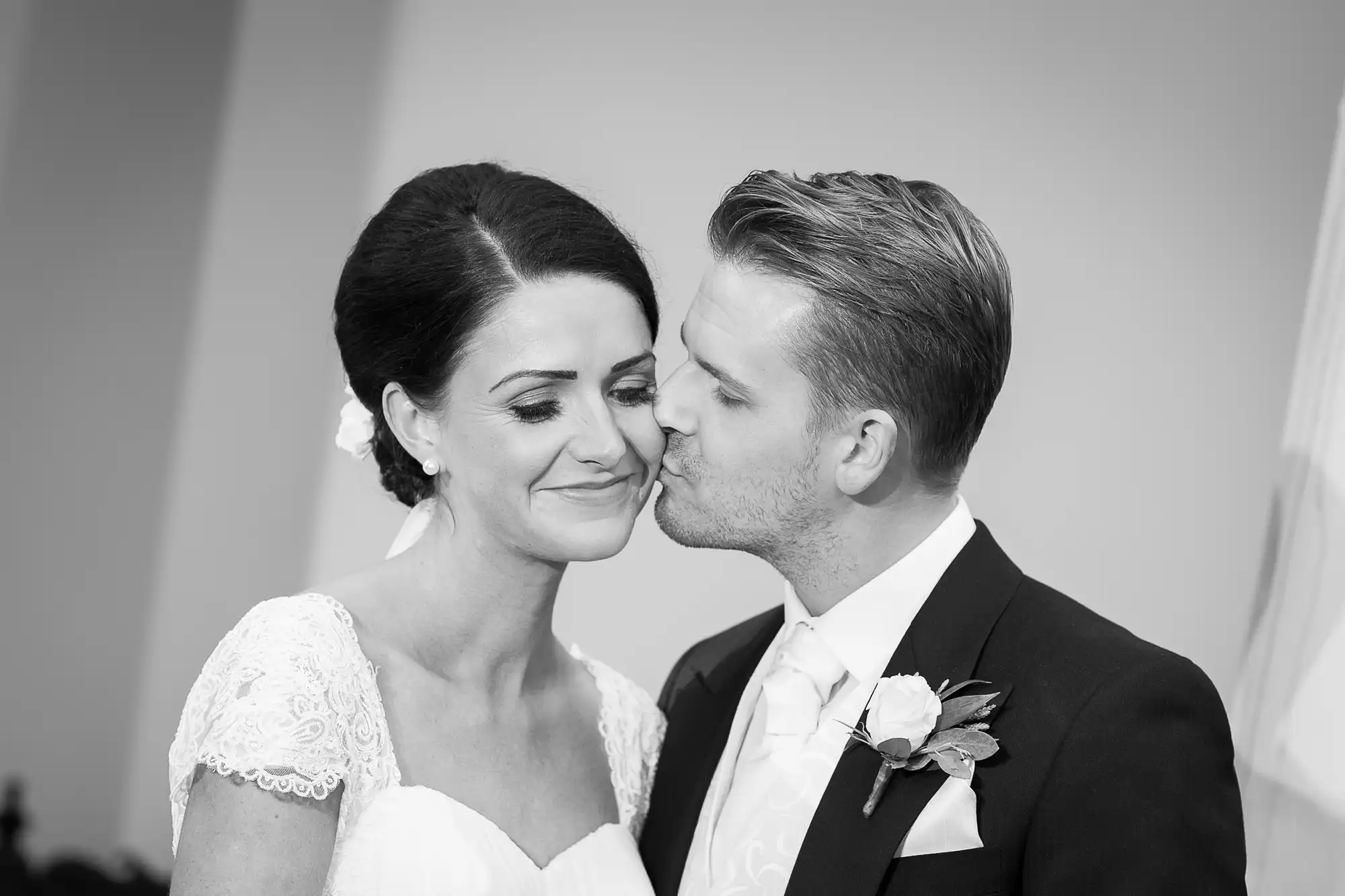 Groom kissing bride on the cheek in a black and white wedding photo, both smiling gently, bride in lace dress.