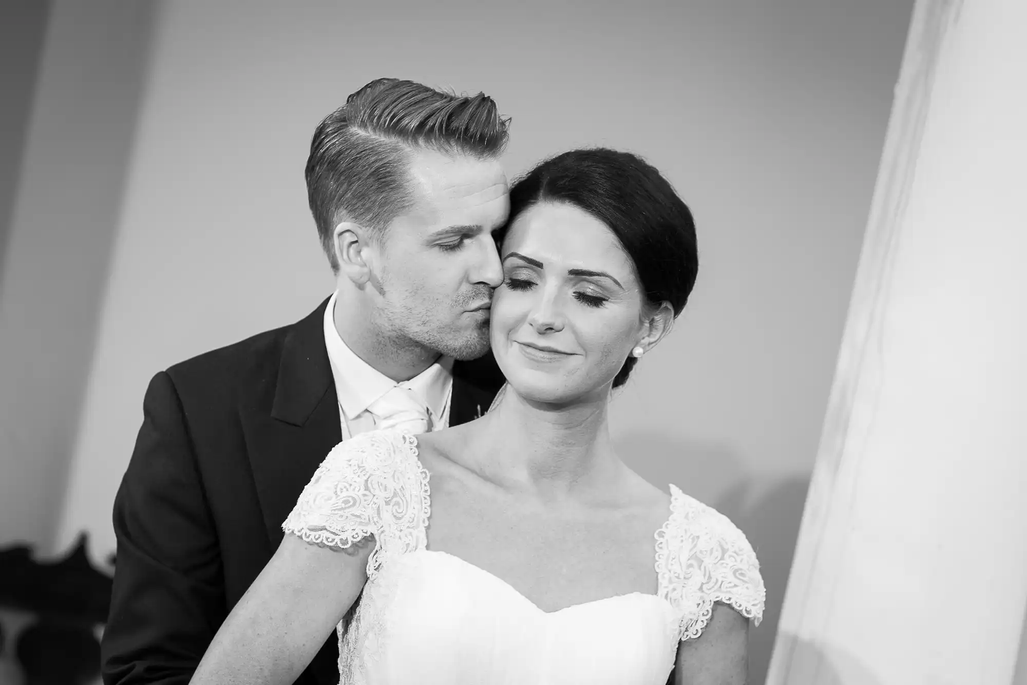 A black and white photo of a groom kissing the bride on the cheek, both dressed in wedding attire with tender expressions.