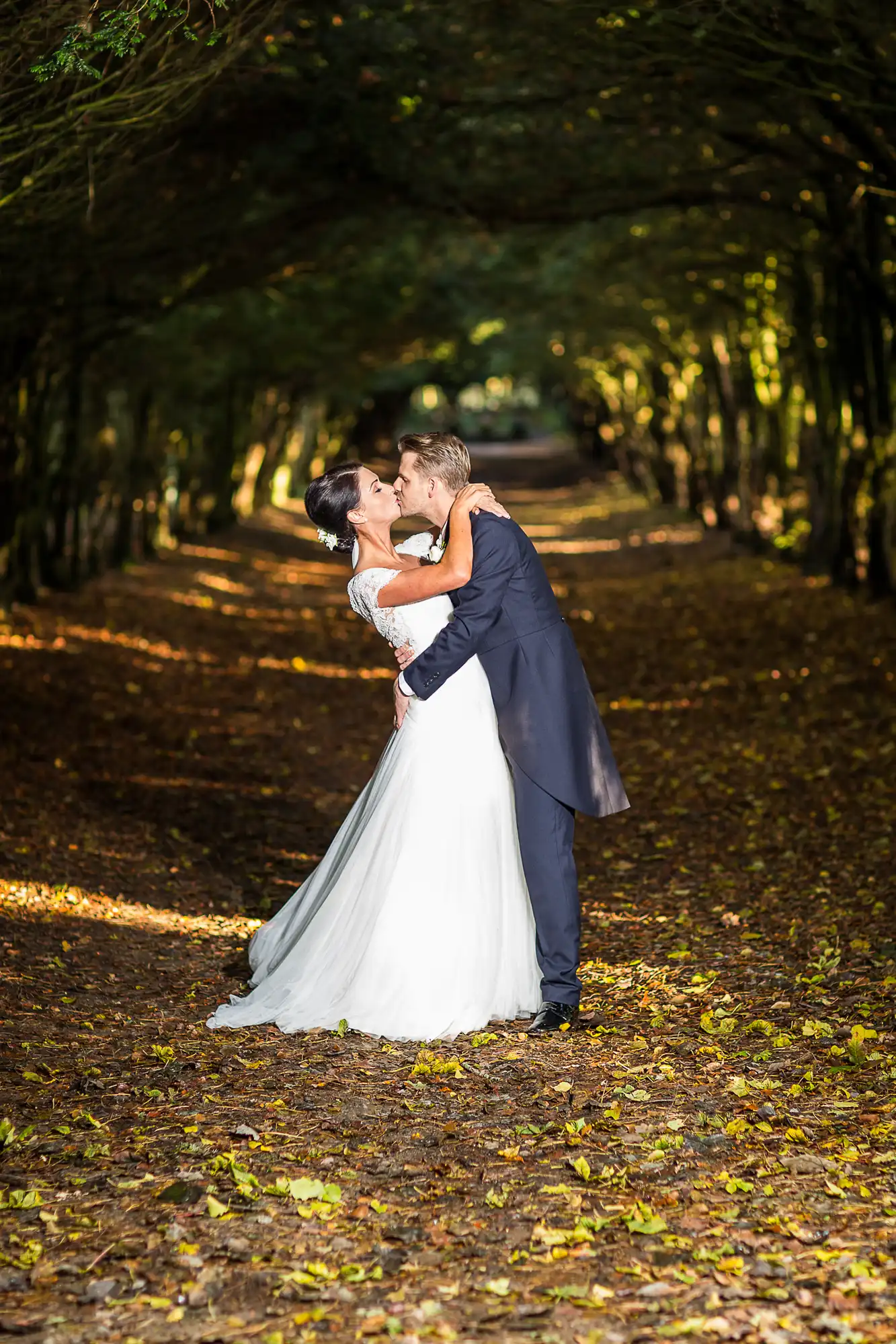 Bride in a white dress and groom in a black suit kissing under a canopy of trees with golden autumn leaves on the ground.