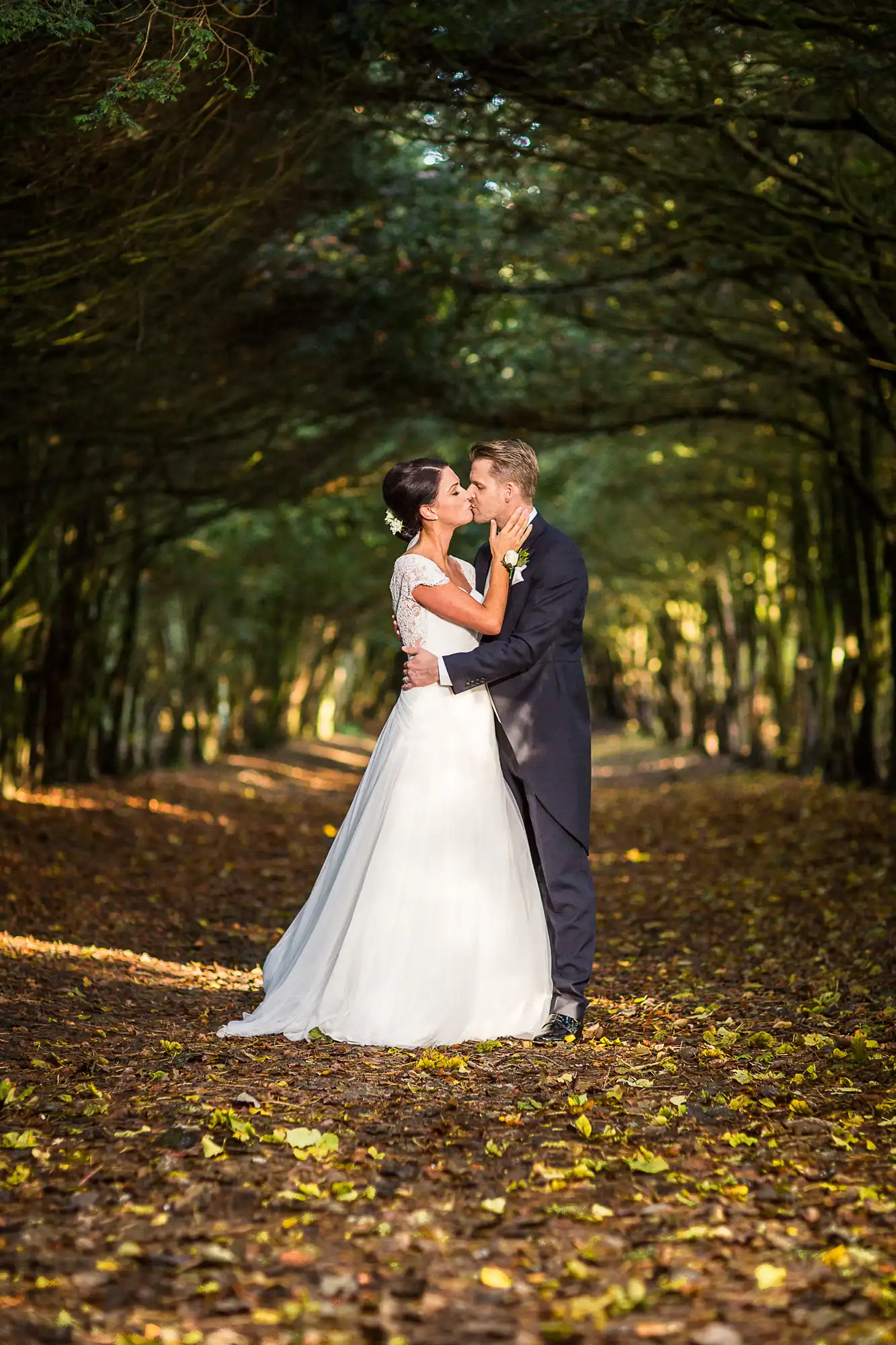 A bride and groom kissing in a forest pathway lined with trees and sunlight filtering through the leaves, with fallen autumn leaves scattered around.