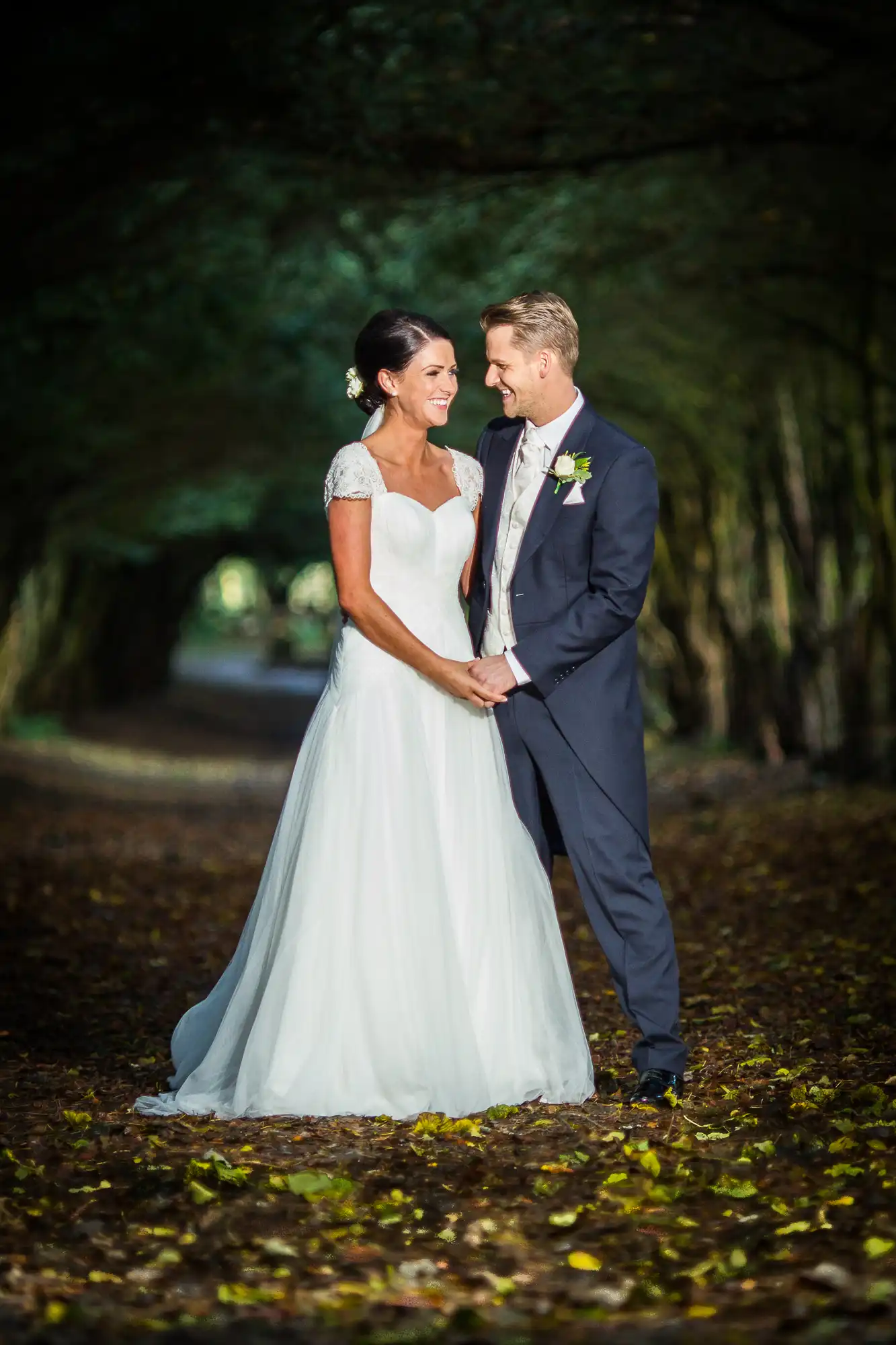 A bride and groom holding hands and smiling at each other on a forest path, surrounded by trees and fallen leaves.