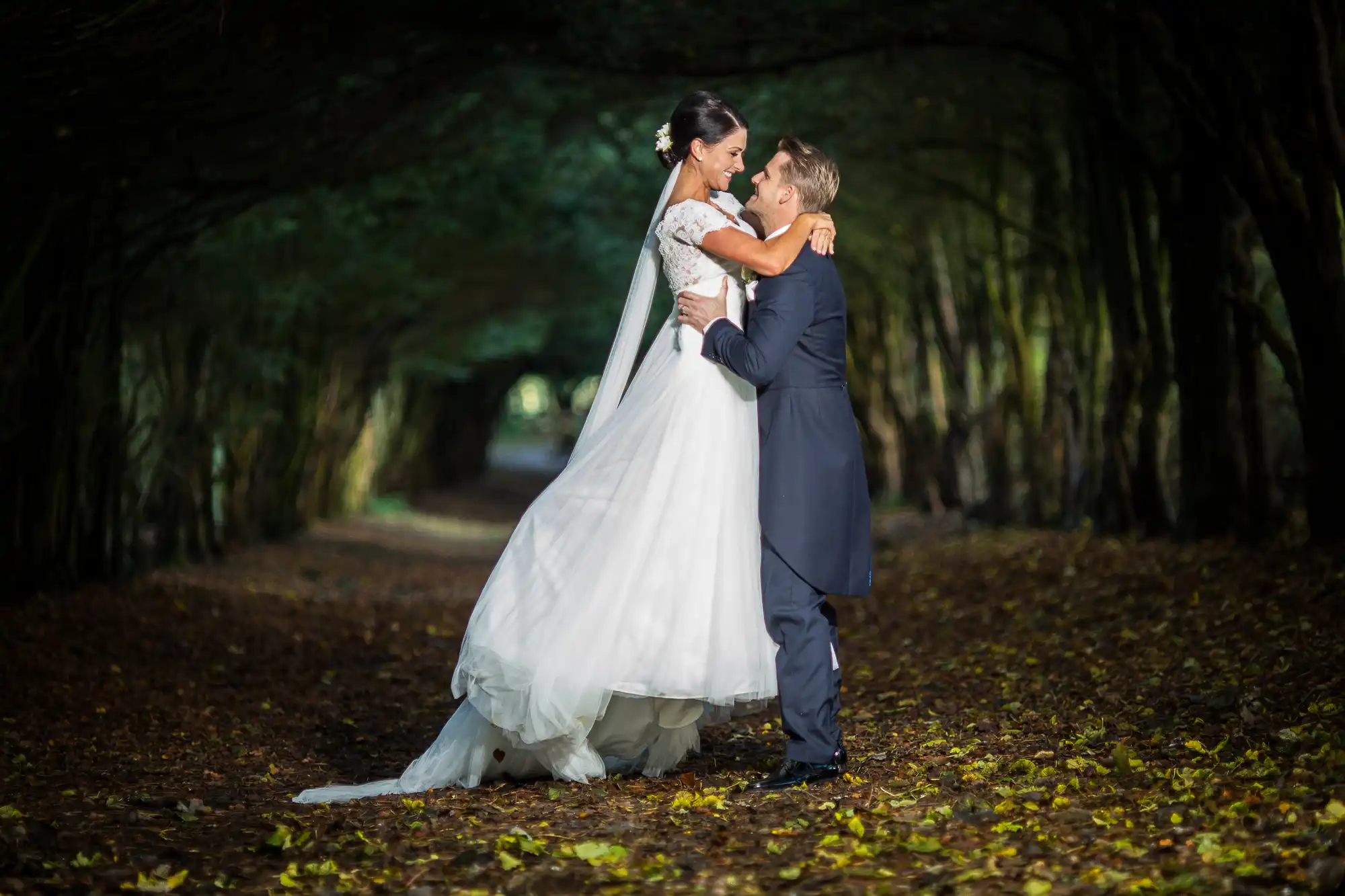 A bride and groom share a loving embrace in a forest pathway, surrounded by trees and fallen leaves, with soft lighting highlighting their joyful expressions.