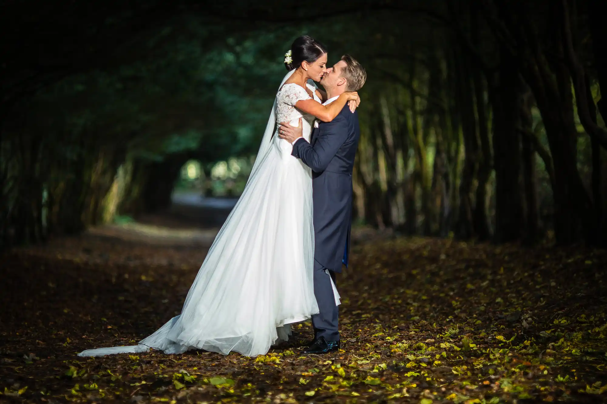 A bride and groom embracing in a tree-lined pathway, sharing a romantic moment, with autumn leaves scattered on the ground.