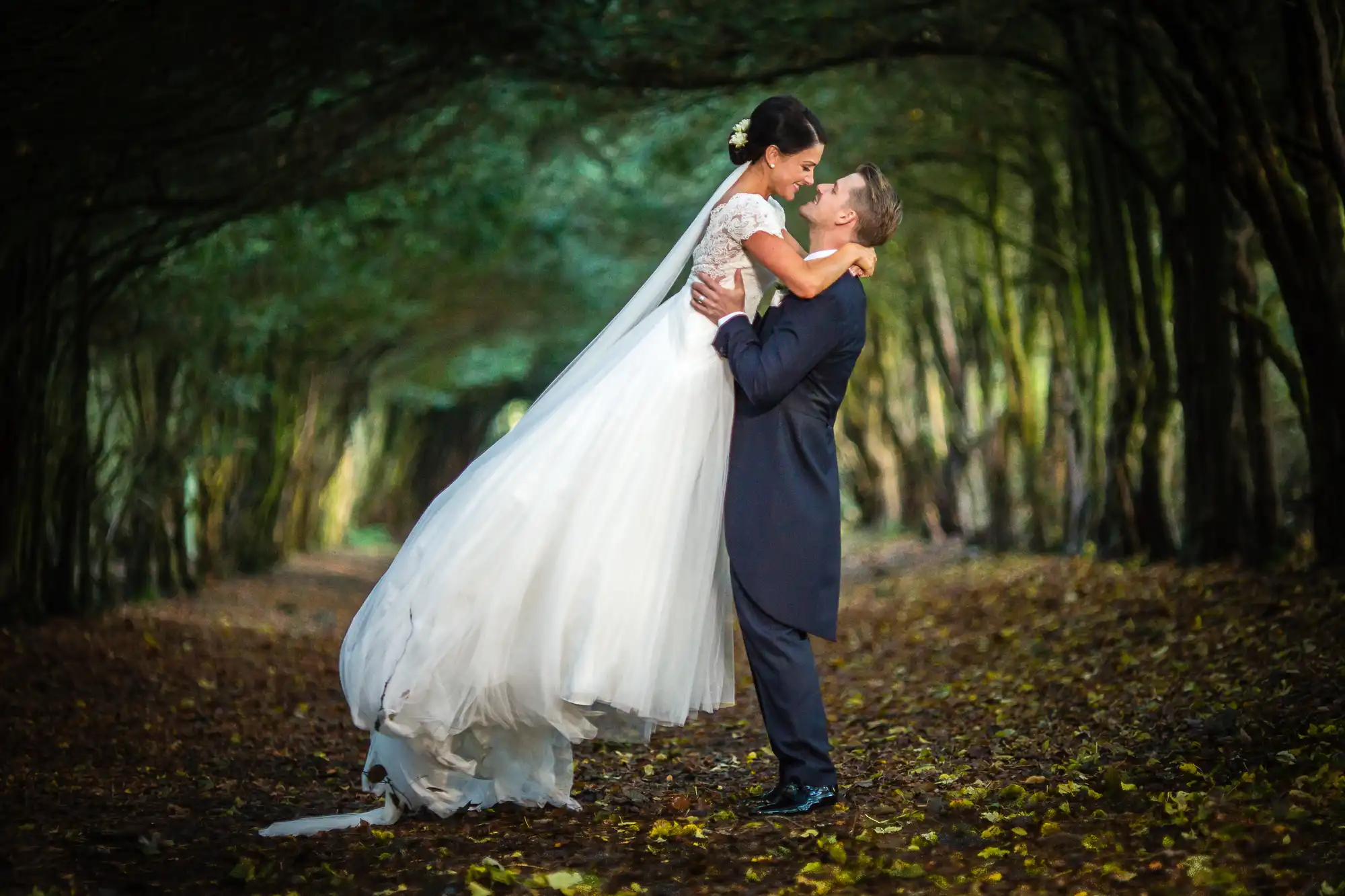 Groom lifting bride in a forested path, kissing under a canopy of trees with autumn leaves on the ground.