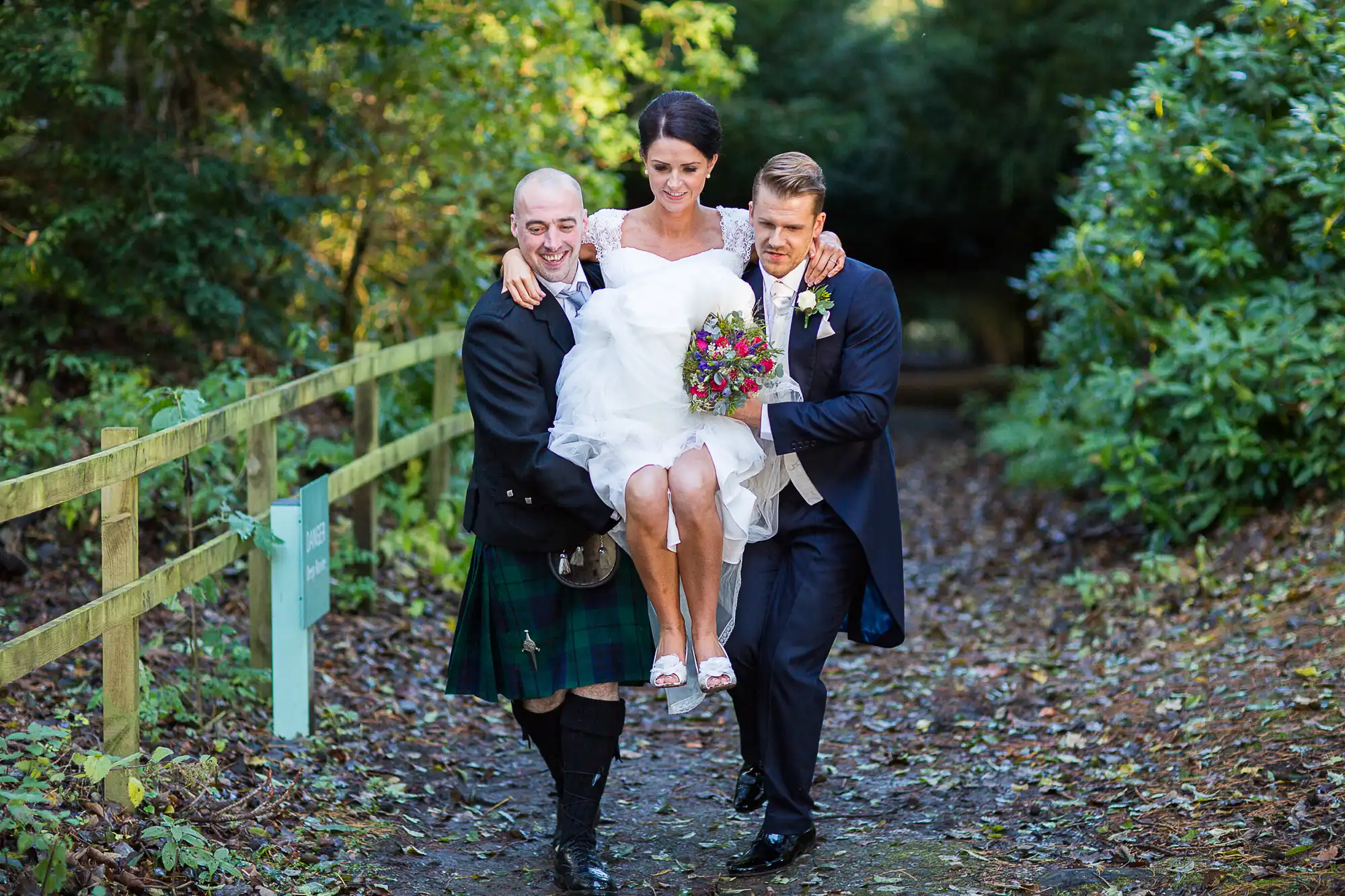A bride being playfully carried by two men, one in a kilt, along a leaf-strewn path with green fencing.