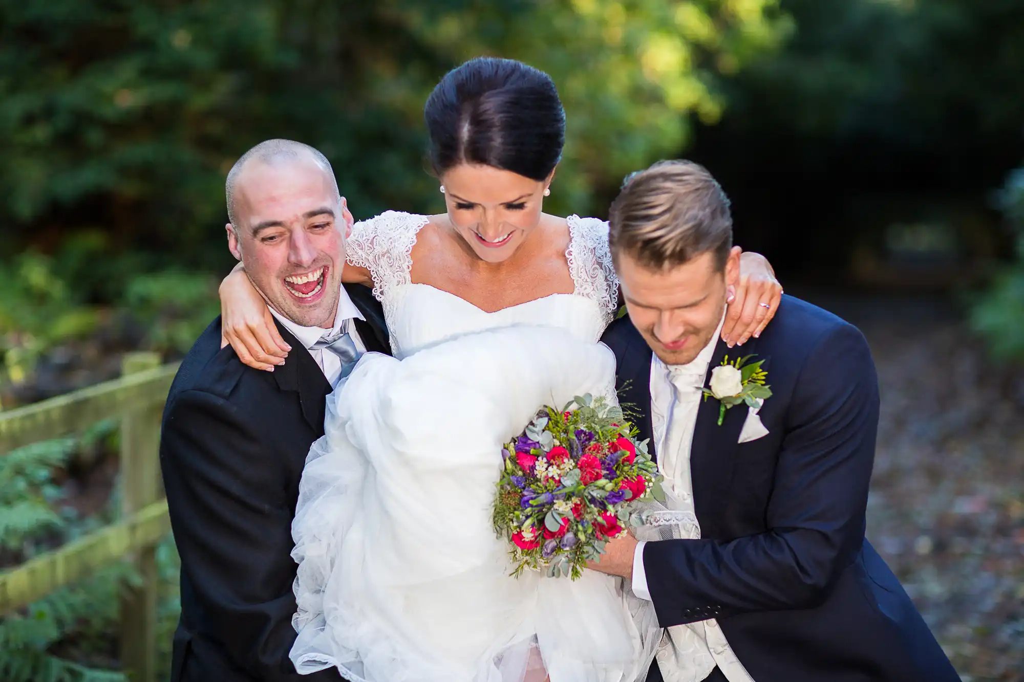 Bride laughing joyfully, flanked by two men in suits, one on either side, all holding a colorful bouquet in a sunlit garden setting.