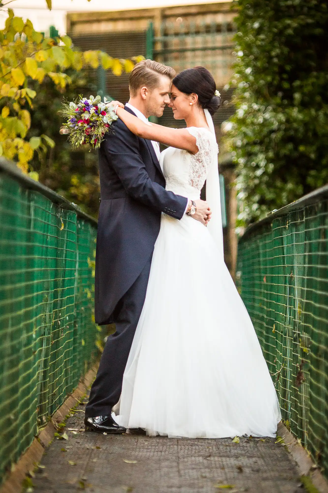 A bride and groom embrace on a narrow pathway, surrounded by green fences, the bride holding a colorful bouquet.