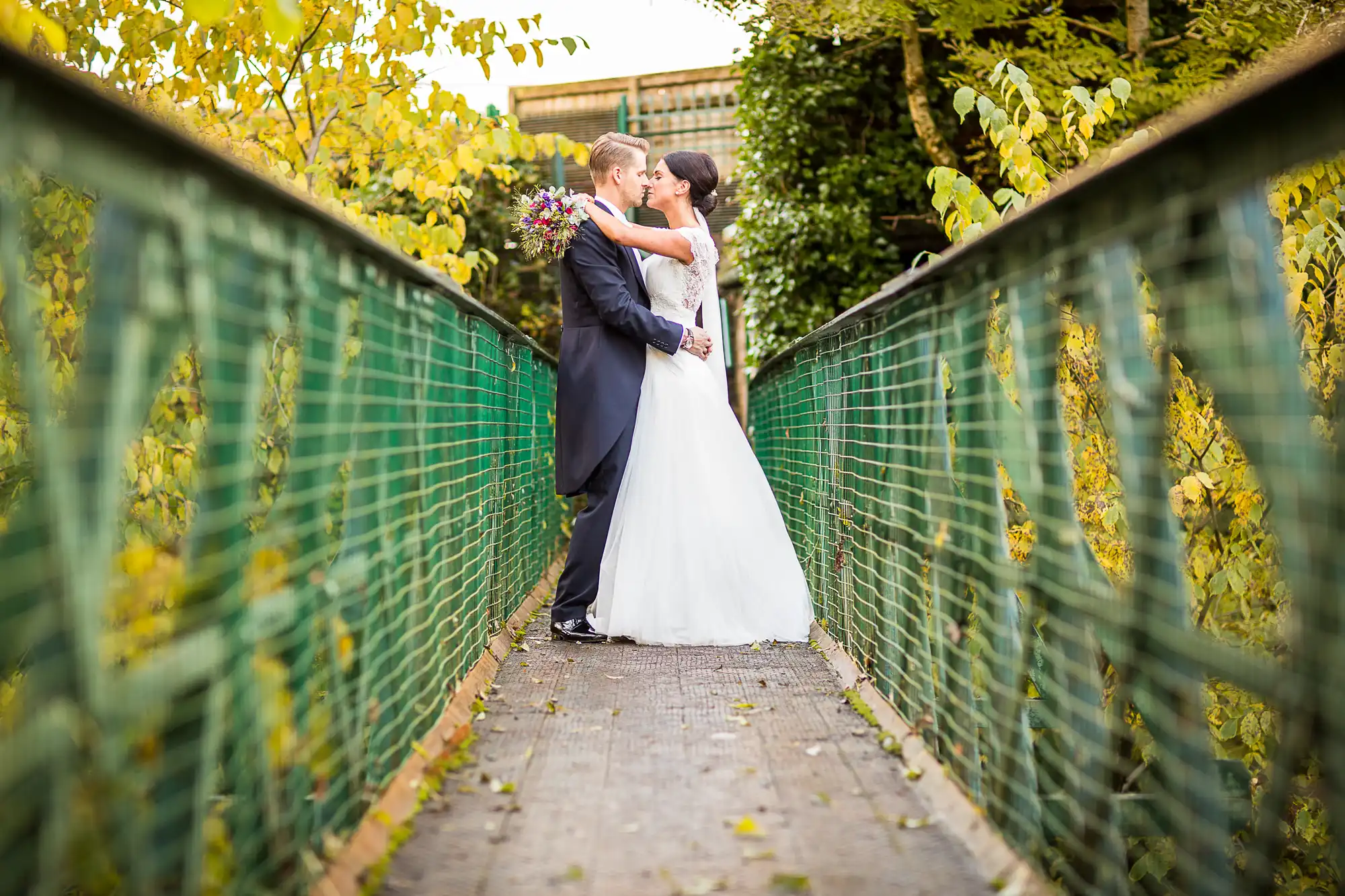 A bride and groom kissing on a bridge surrounded by green mesh and autumn leaves.